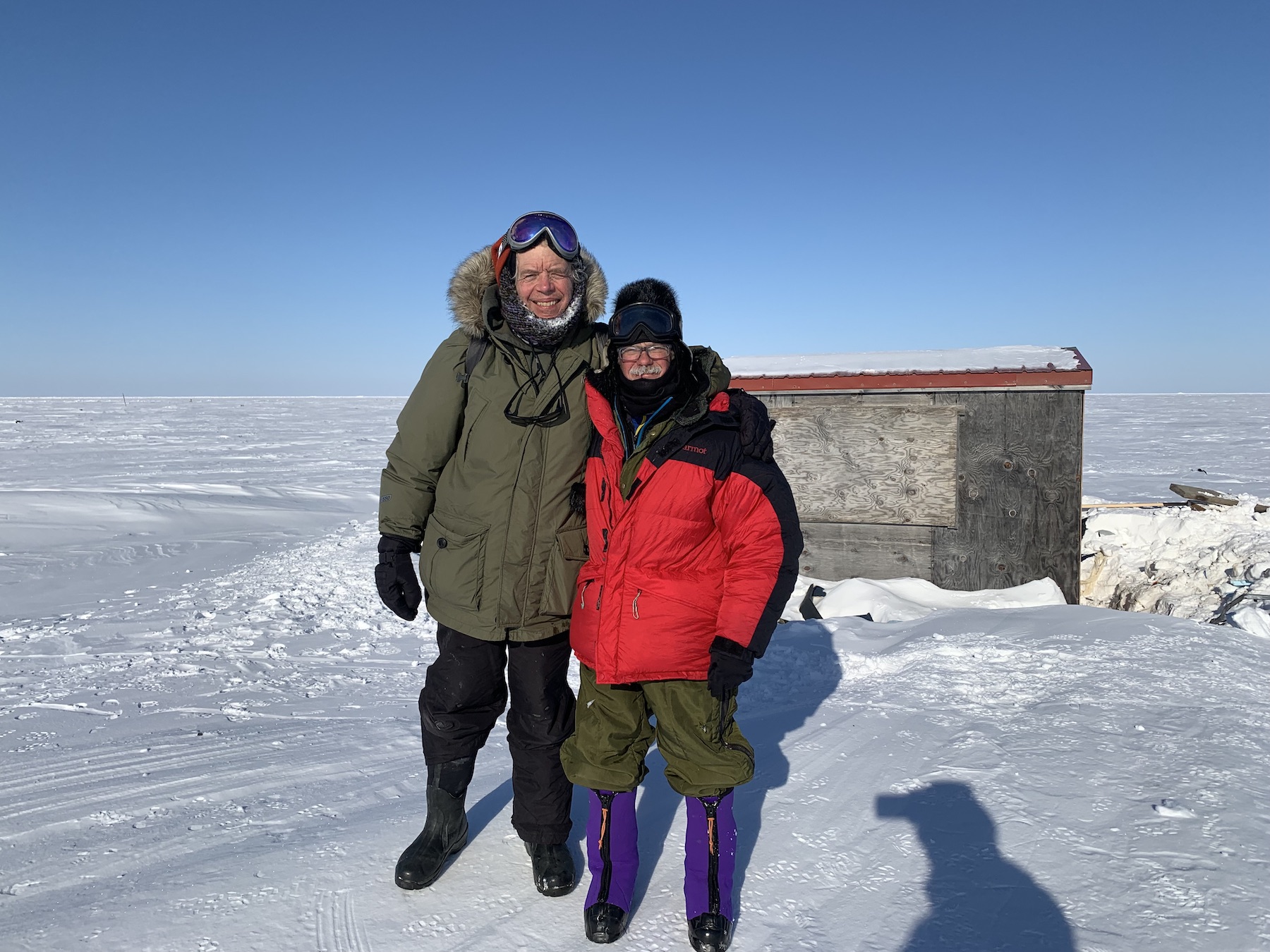 In bright sun, two men in winter gear pose in front of a plywood structure in a wide plain of snow stretching out across sea ice to the horizon.