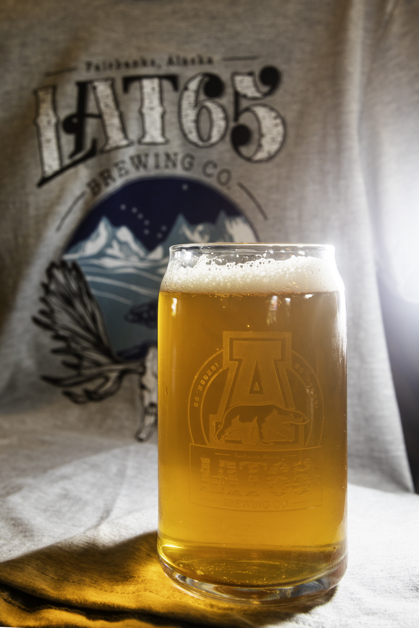 A glass of golden beer in front of a gray shirt with a brewery logo on it.