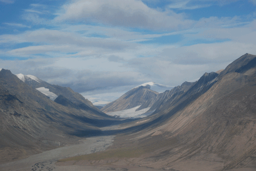 A parabola-shaped valley between tundra-covered mountains features a braided river draining from a glacier.