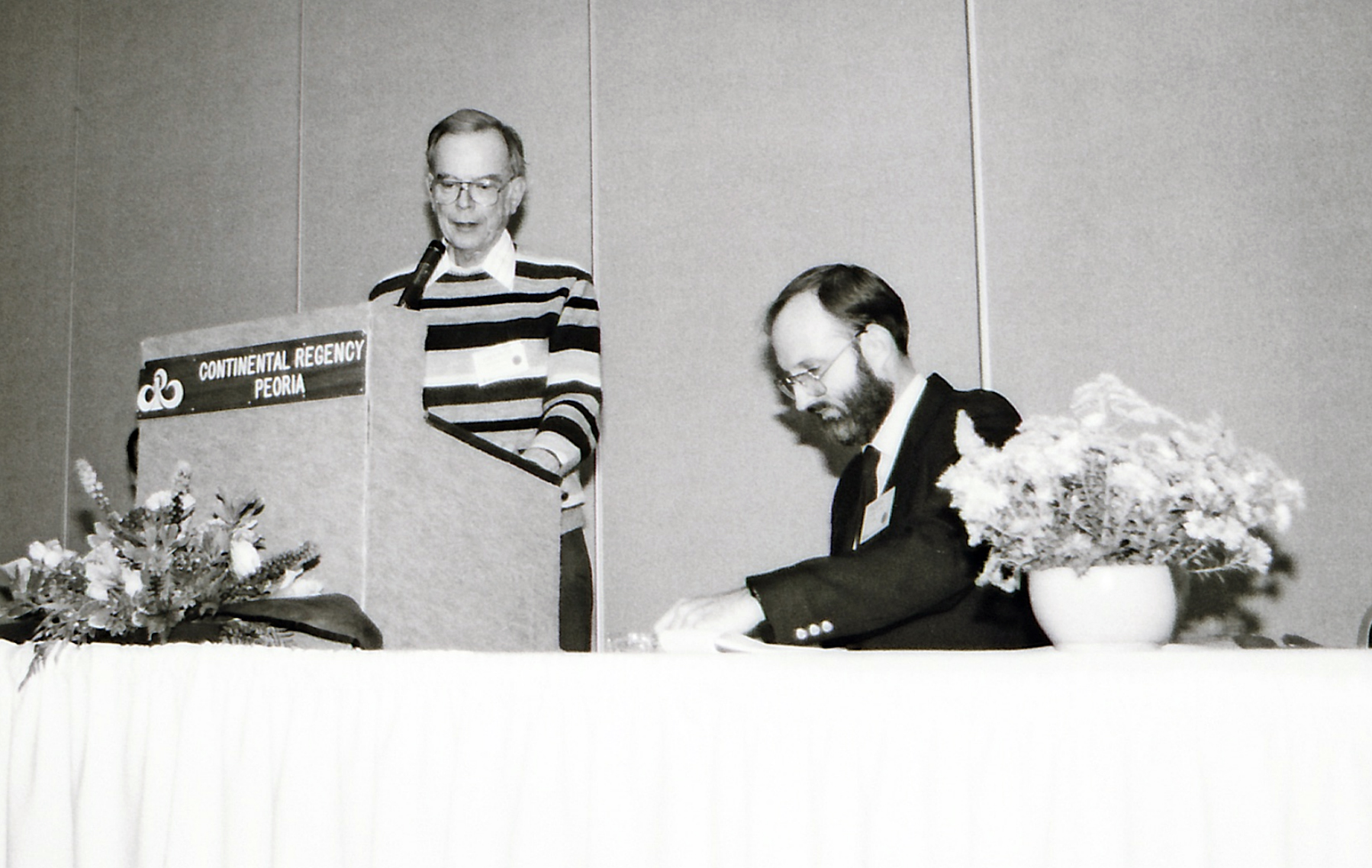 In this black and white picture, a man stands at a podium while another sits looking at material on a nearby table 