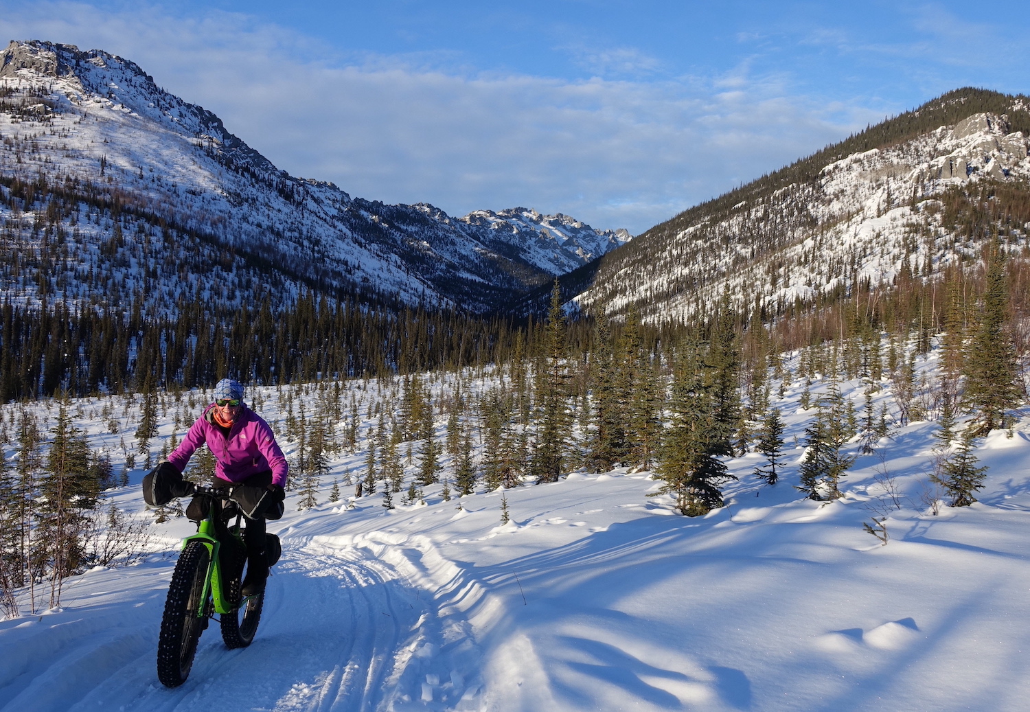 A woman rides a fat-tired bike on a snow trail surrounded by small spruce trees in a valley between mountains.