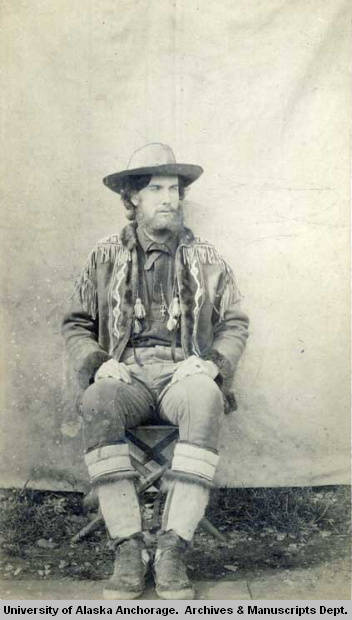 A bearded young man dressed in fringed leather clothing, skin boots and a broad-brimmed hat sits in front of a white canvas backdrop.