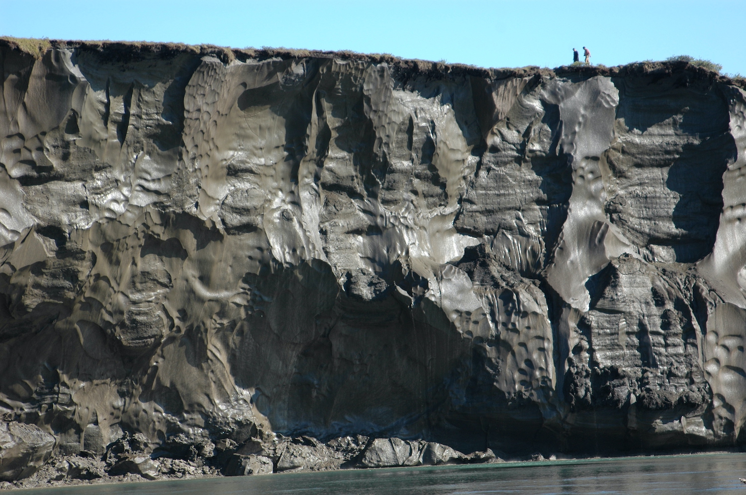 A frozen wall of icy mud rises above a water body. Two people walk along the top of the wall.