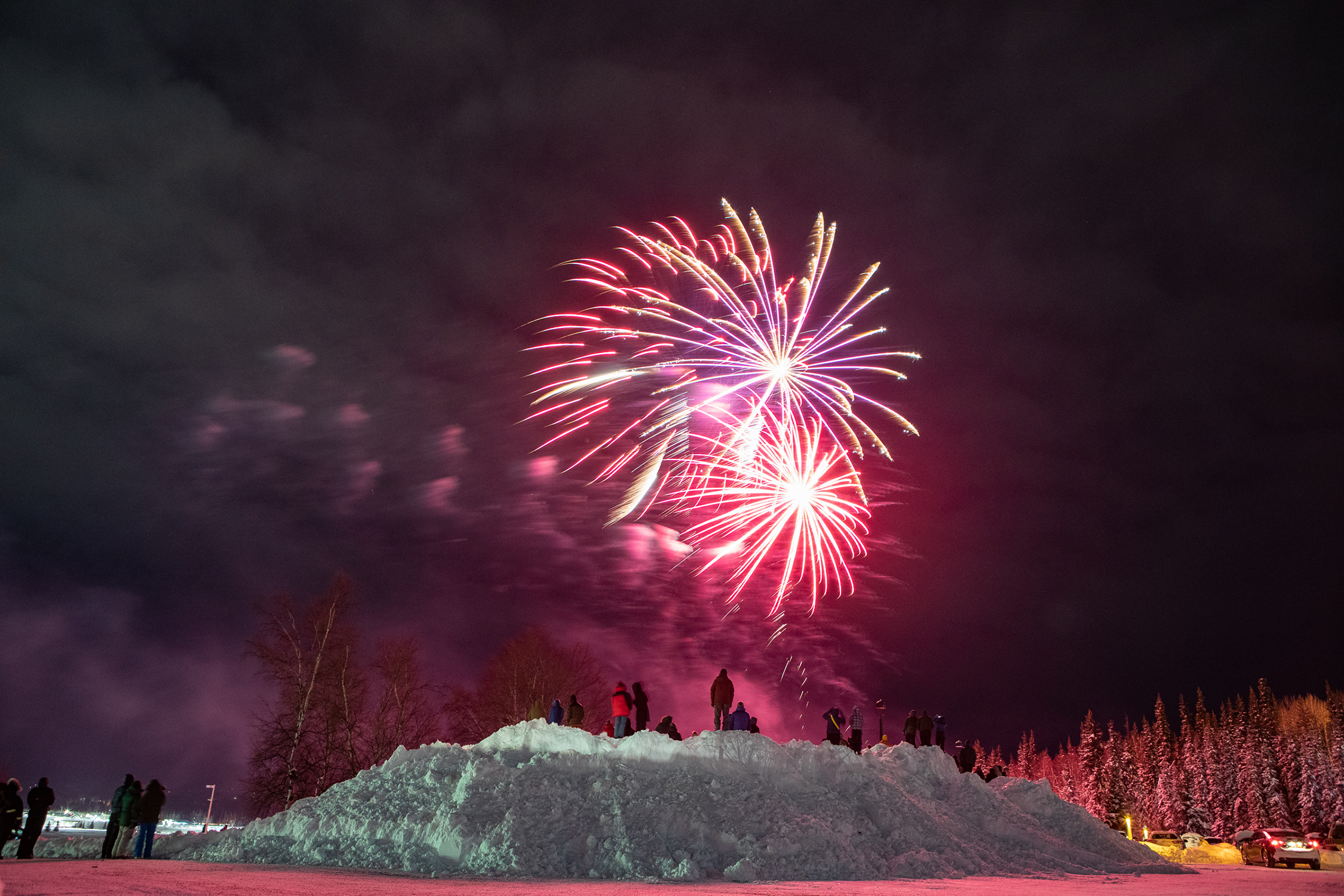 people stand on a snow pile watching fireworks in the sky
