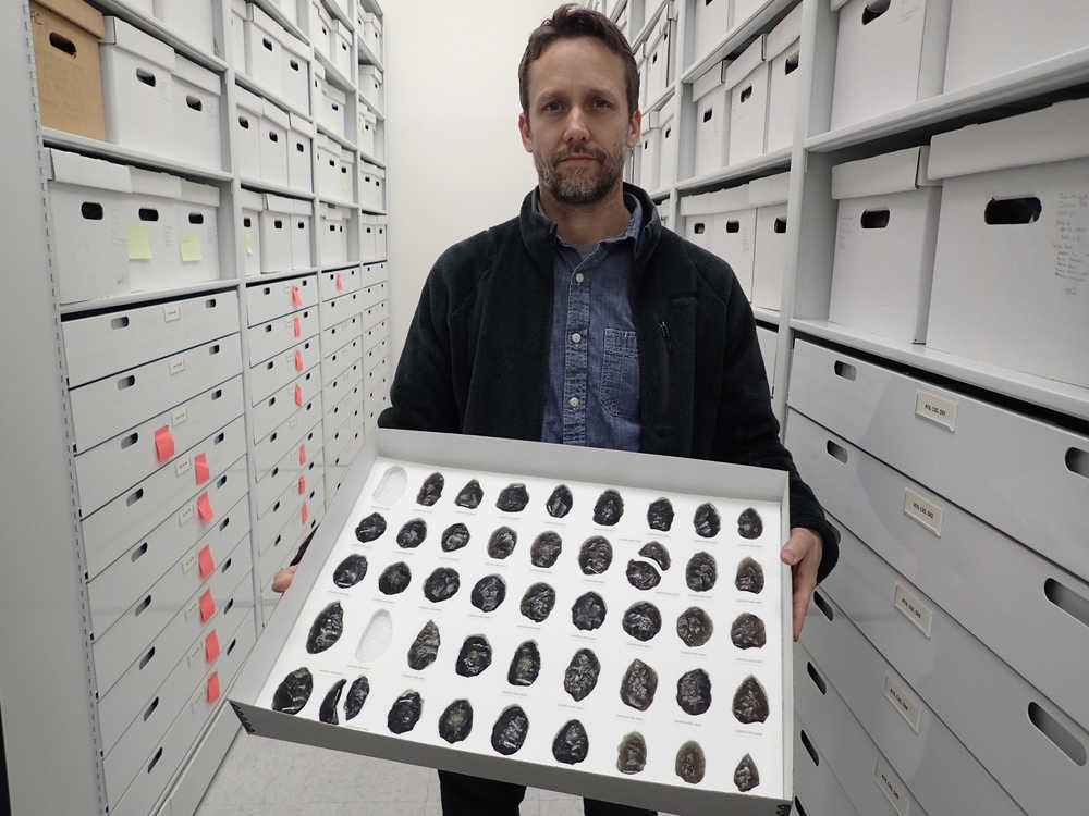A man stands between two rows of white shelving while holding a tray with about 40 shiny black rocks held in holes cut to fit from white foam.
