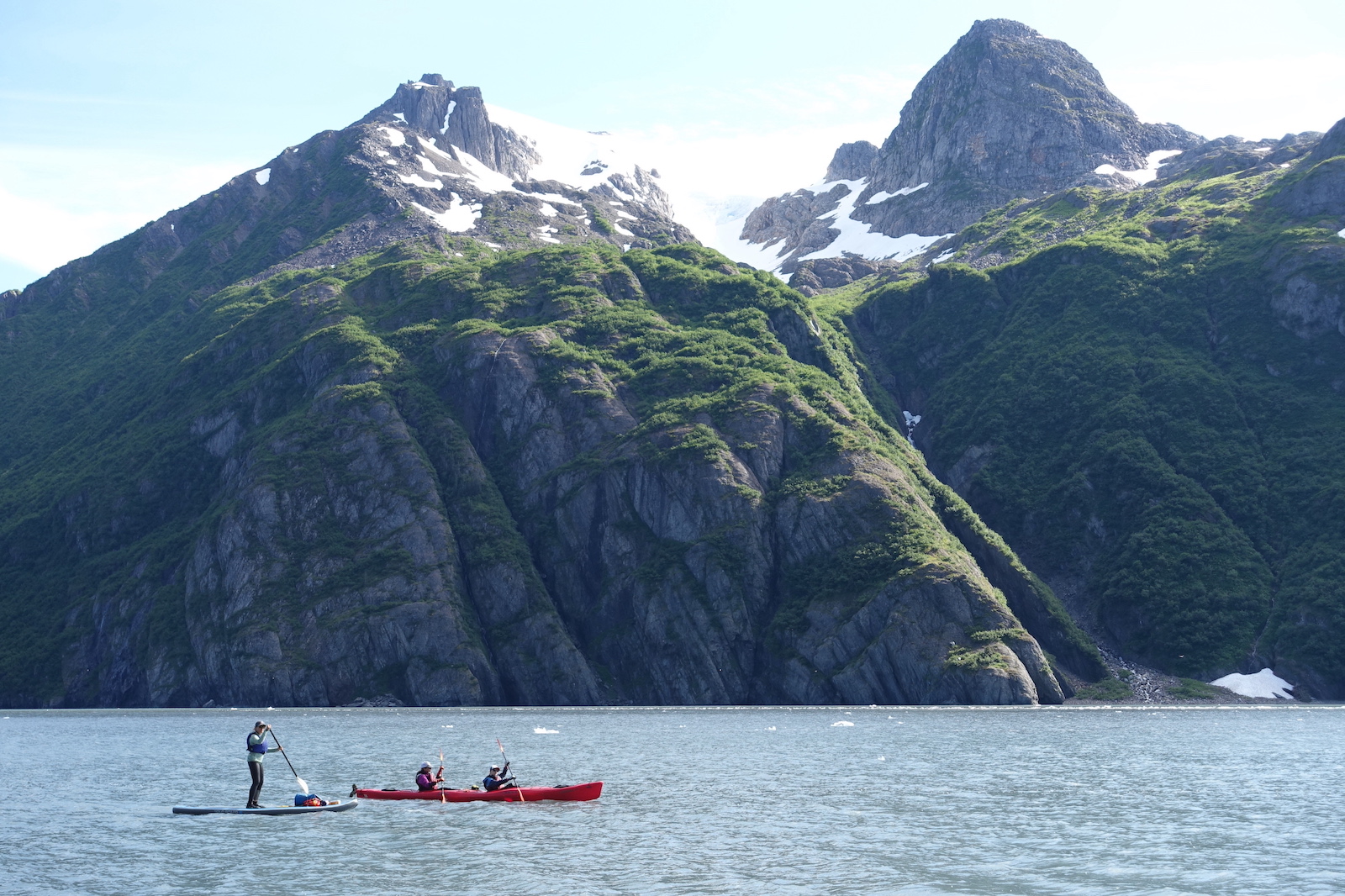 A paddle boarder and two people in a tandem kayak paddle on a water body with snow-laced mountains and high cliffs topped with green in the background.