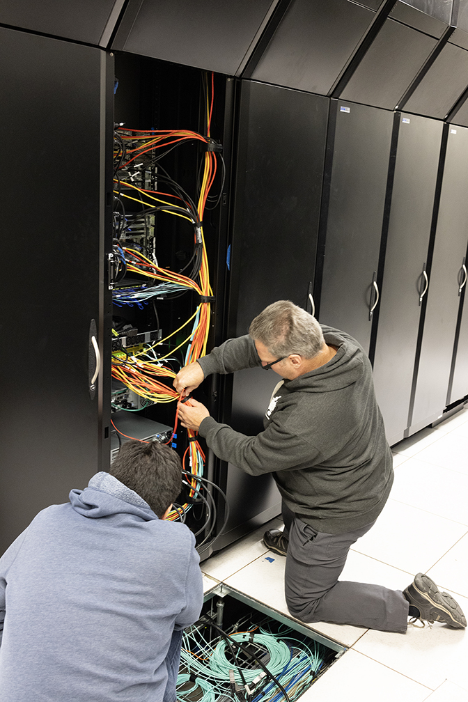 Two men kneel to work on wiring in an open computer cabinet, one of a long wall of closed black cabinets.