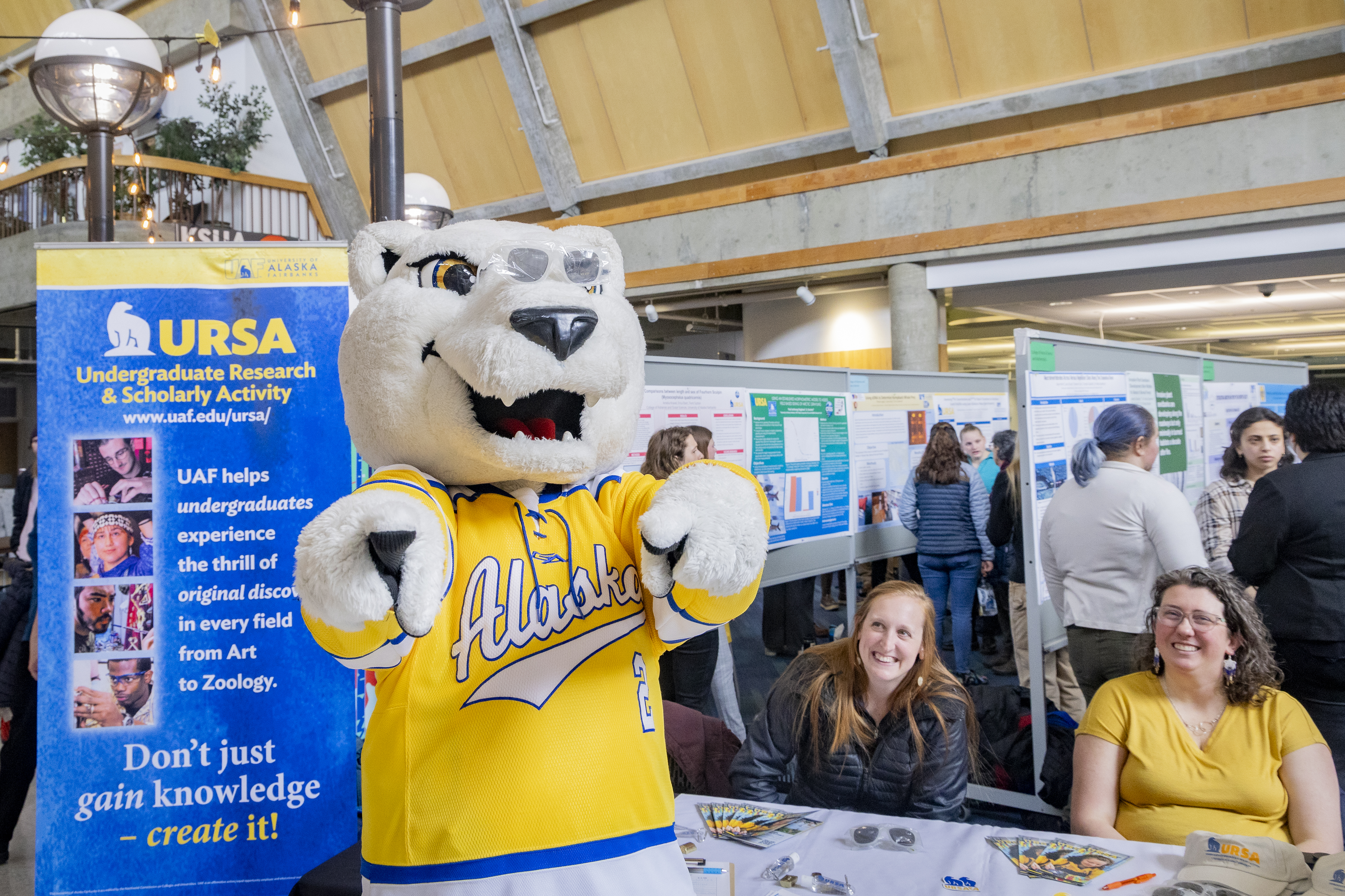 A mascot bear stands next to a table where two people are sitting.