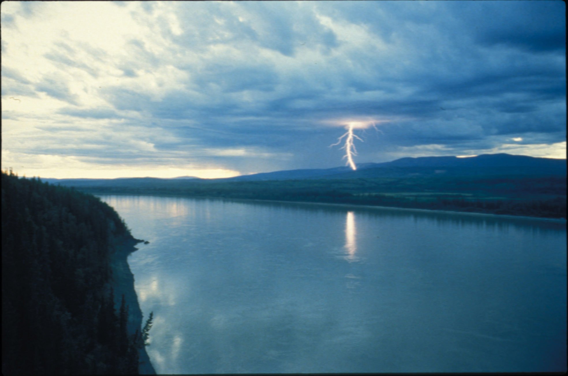 A lightning bolt strikes a green forested hillside beyond a wide river in the foreground.