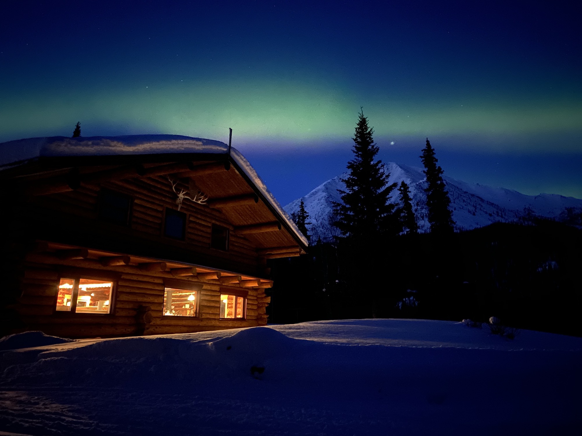 Lights from inside a log building illuminate snow in the foreground at night. A clear sky features a green aurora borealis. A snowy mountain rises in the background.