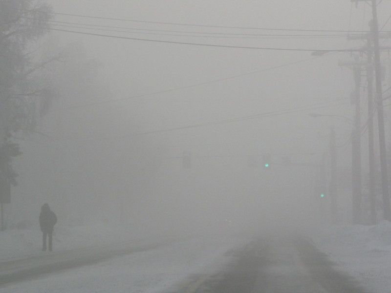  A man walks through thick fog along a roadway. Green traffic lights are barely visible in the background.
