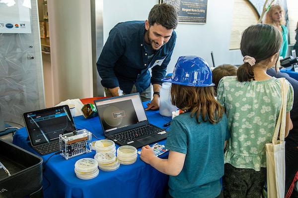 Scientist talks to two young children at a table with petri dishes and a computer.