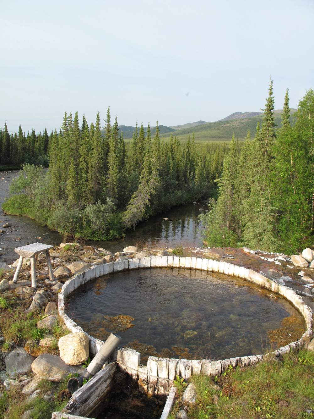 A pool of water hemmed in by a circle of upright boards overlooks a shallow clear river flowing through a spruce forest with tundra-covered hills in the background.