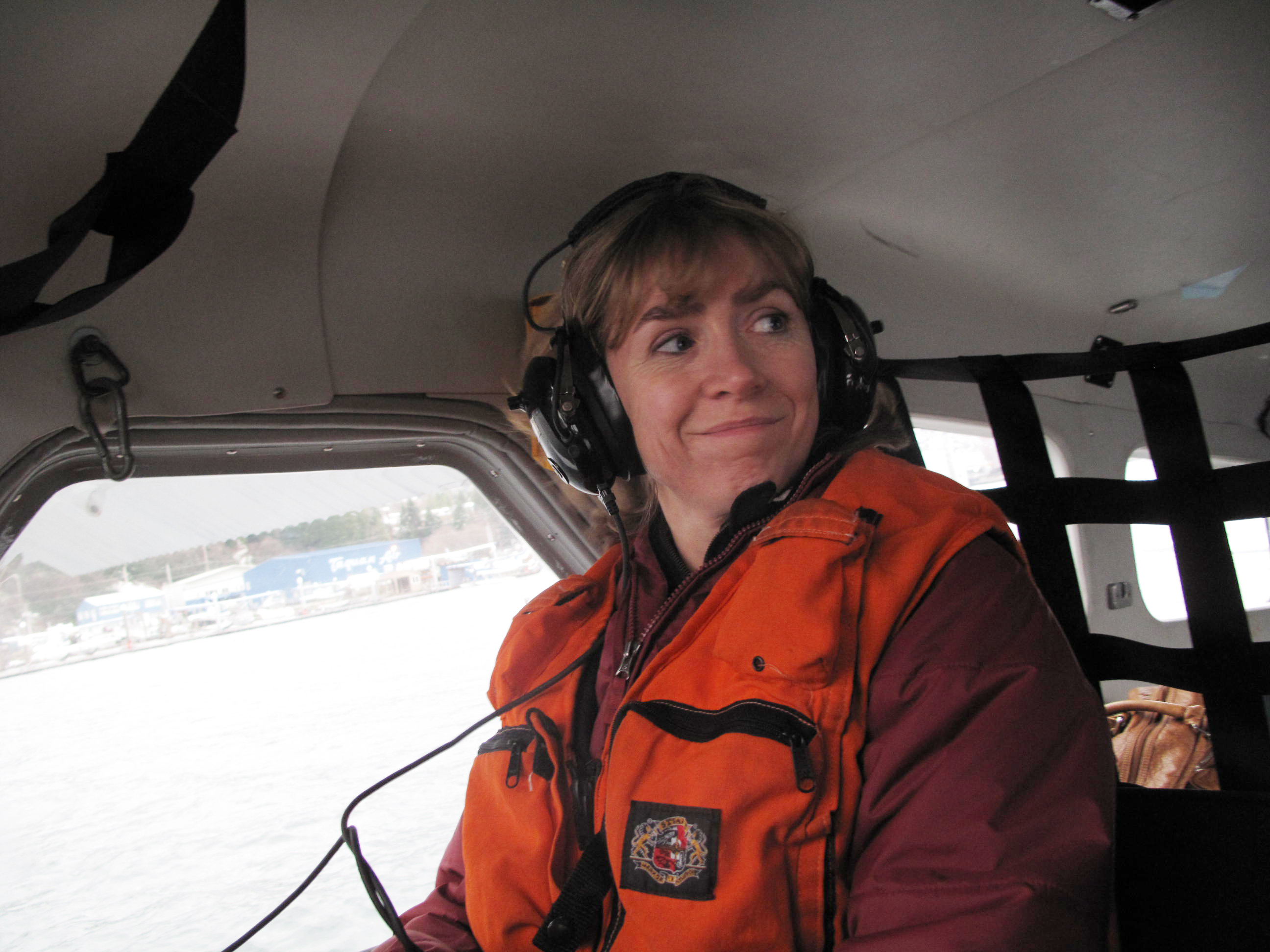 A woman wearing an orange vest and earphones smiles while sitting in a small aircraft.