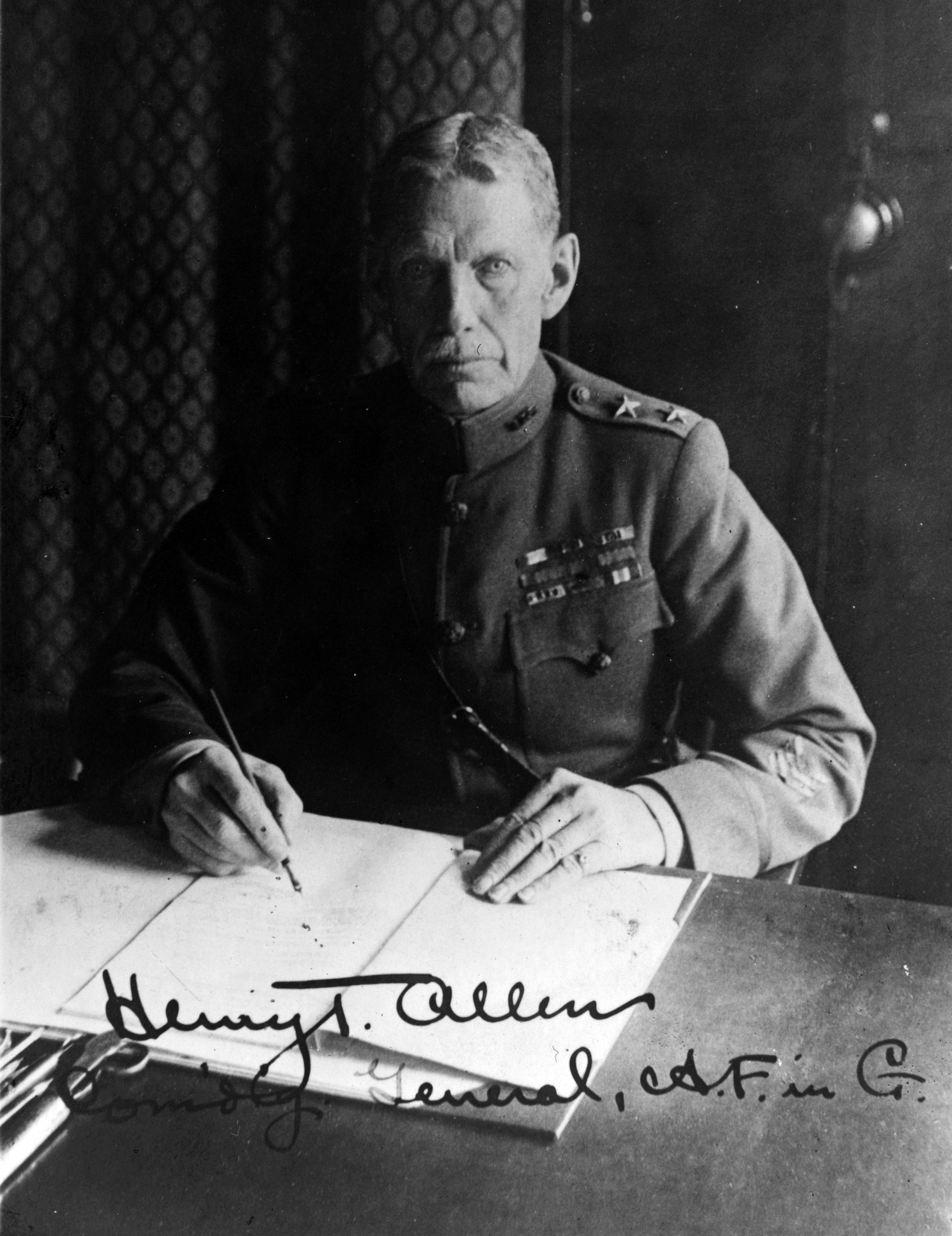 A man in military uniform sits at a desk and writes with a fountain pen in a black and white photograph signed "Henry T. Allen, Com'di'g General, A.F. in G."