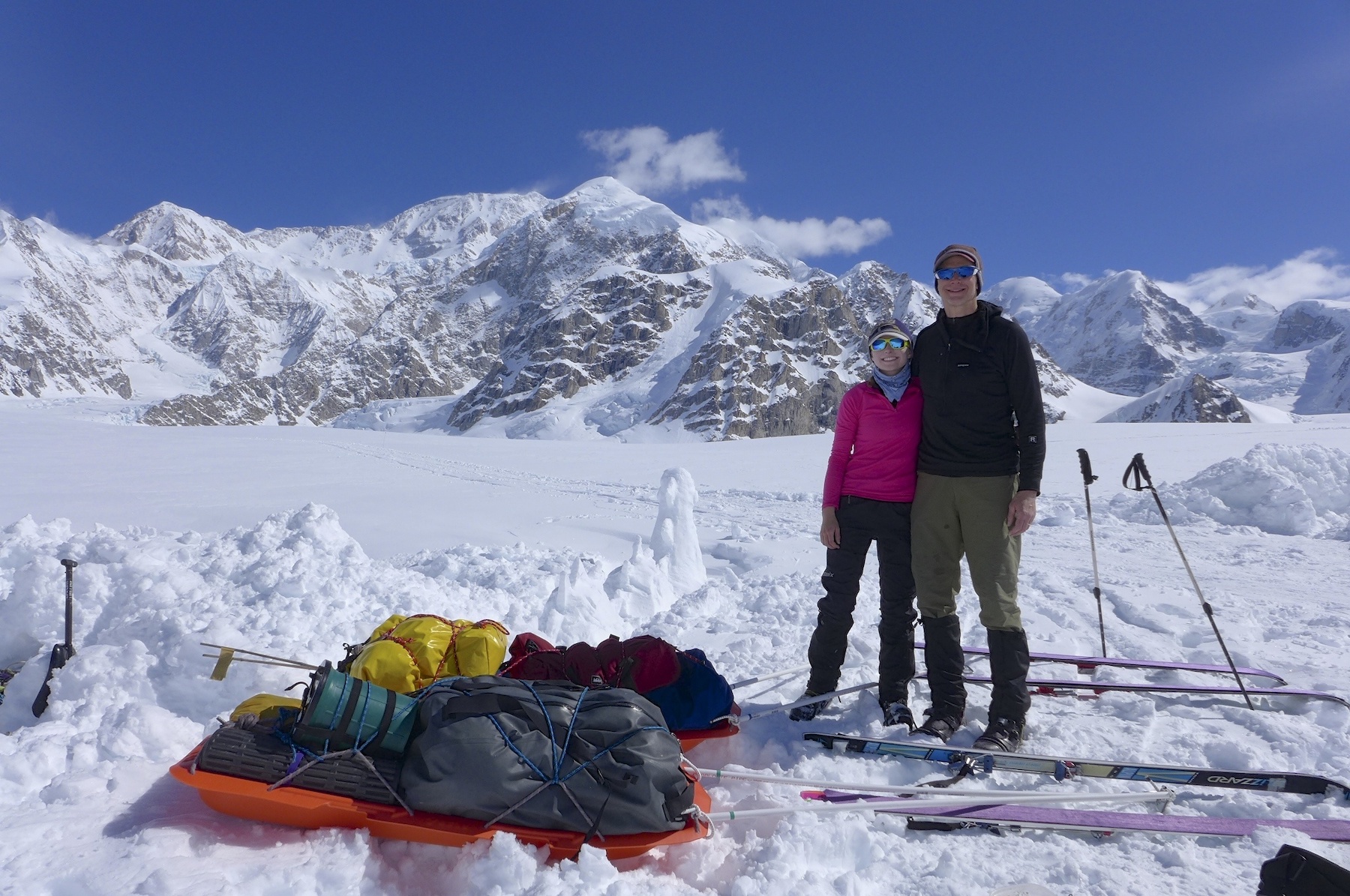 A man and his daughter stand together on a snow-covered glacier with snowy and rocky mountains in the background.