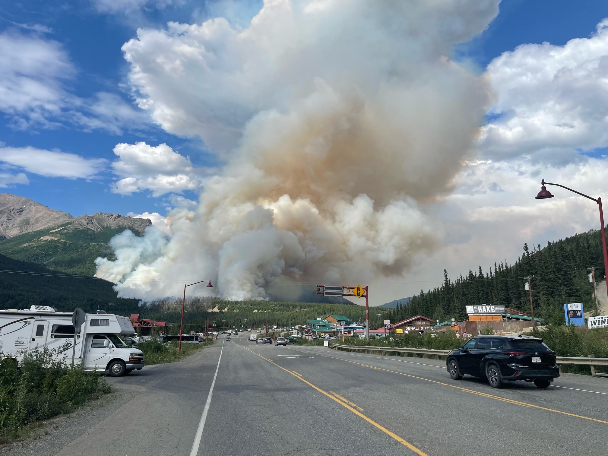 In between mountains, a broad column of smoke rises from a evergreen forest just beyond a highway lined with buildings.