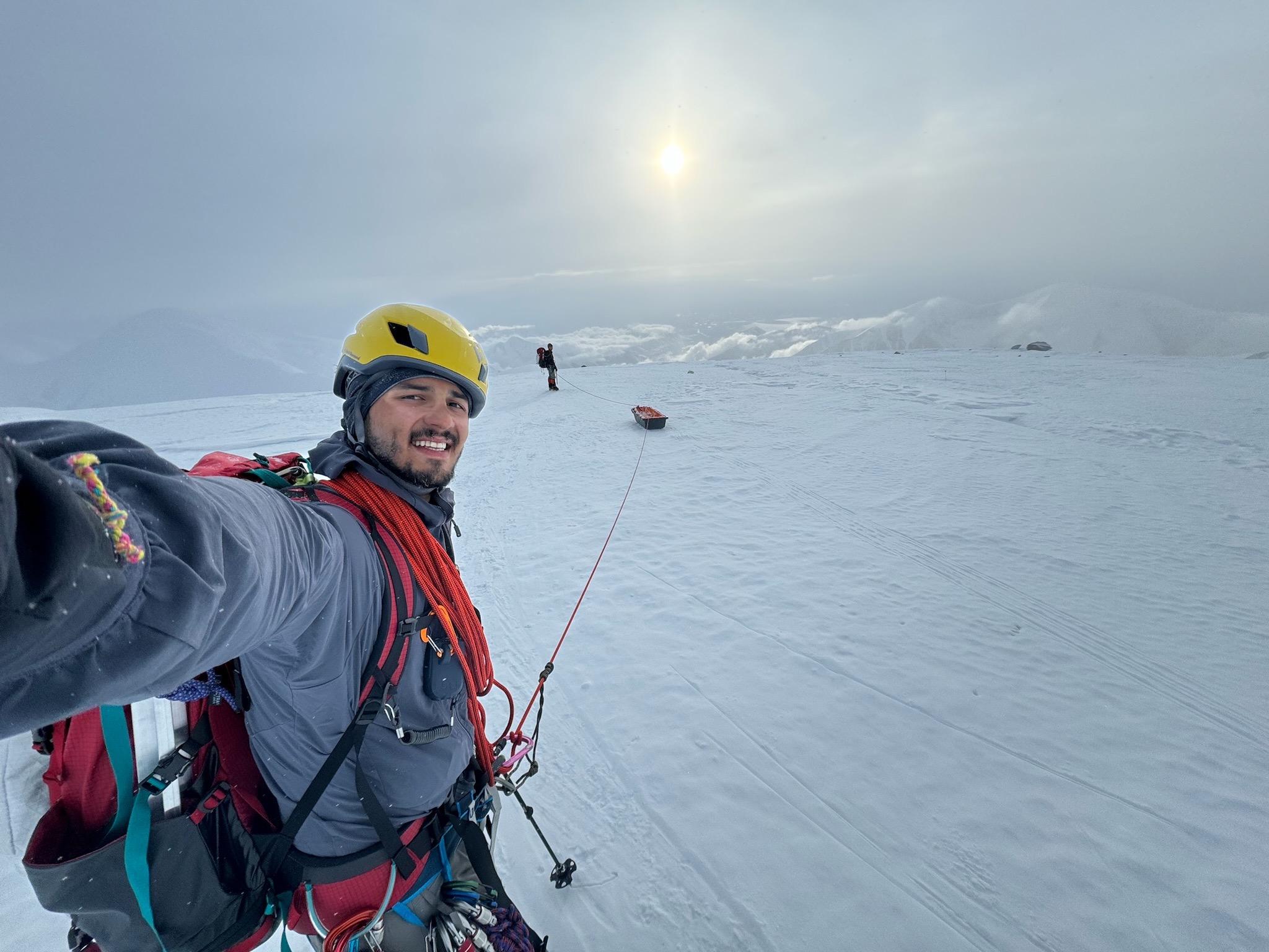 A man takes a photo of himself on a snowy mountain slope. He is roped to a sled and another man in the background.