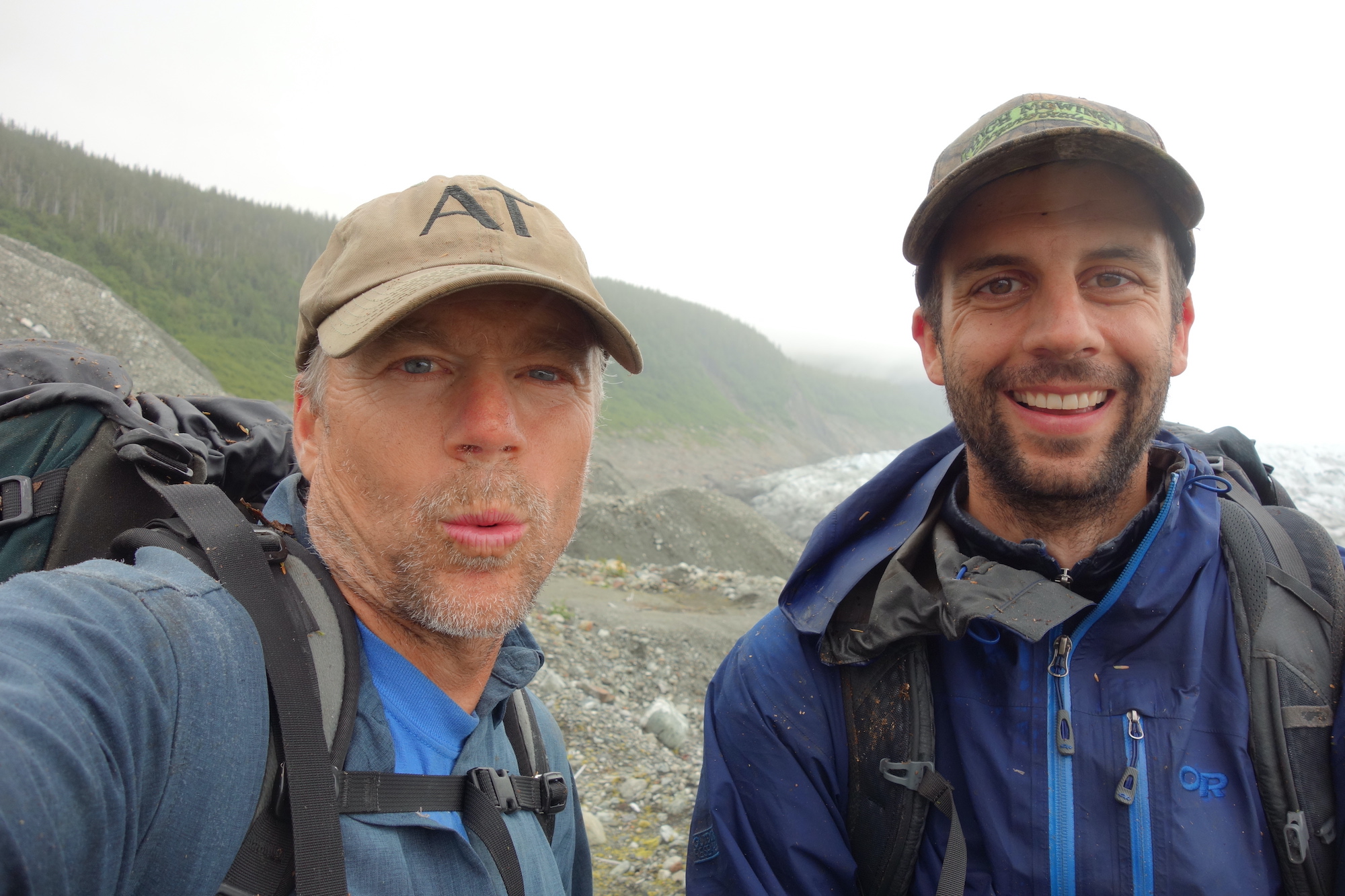 Two men smile at the camera while standing in a gravel-strewn area left by the recent melting of a glacier.