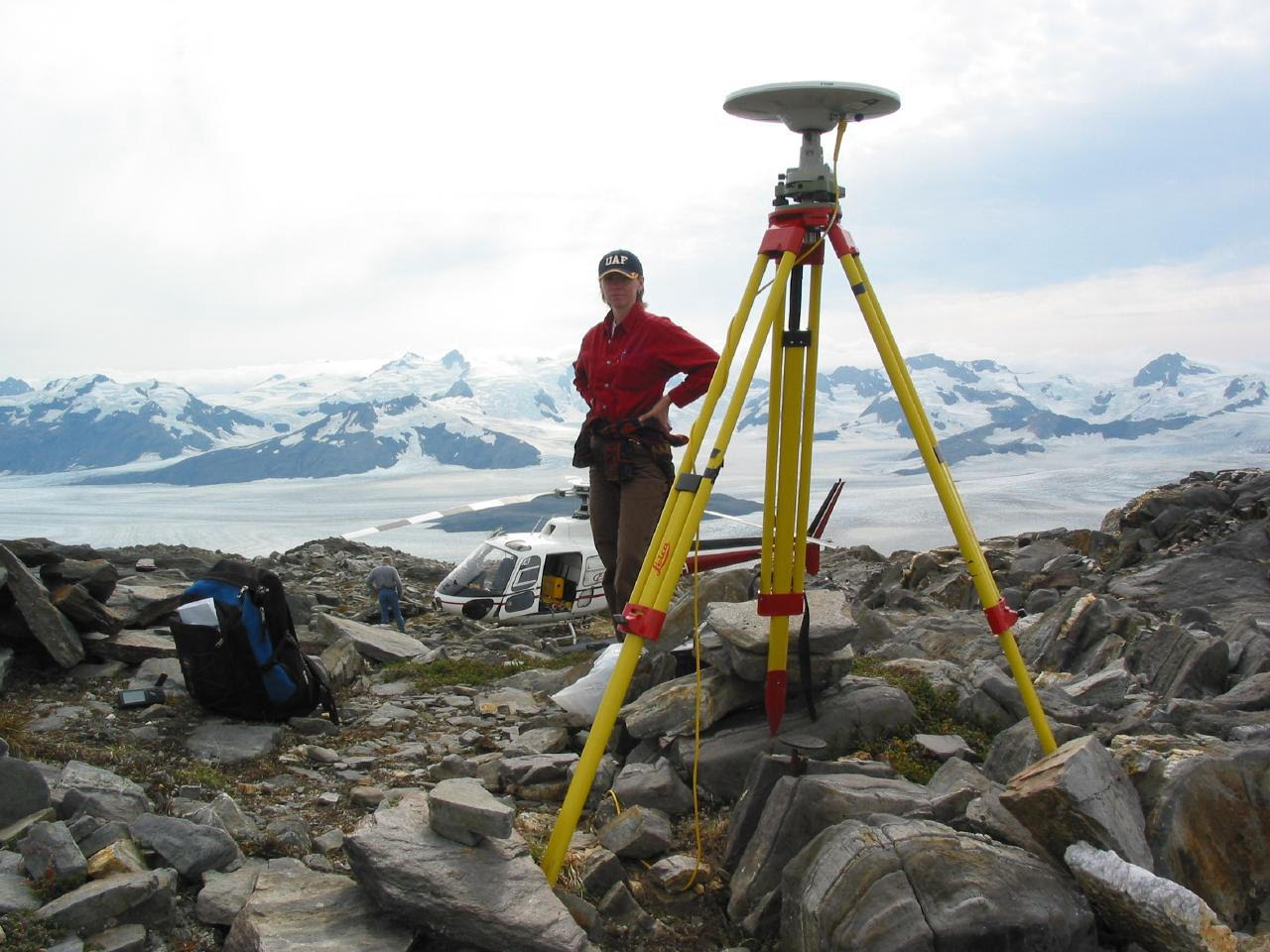 A woman stands on rocky ground with a surveying device on a tripod in the foreground and glacier-covered mountains in the background.