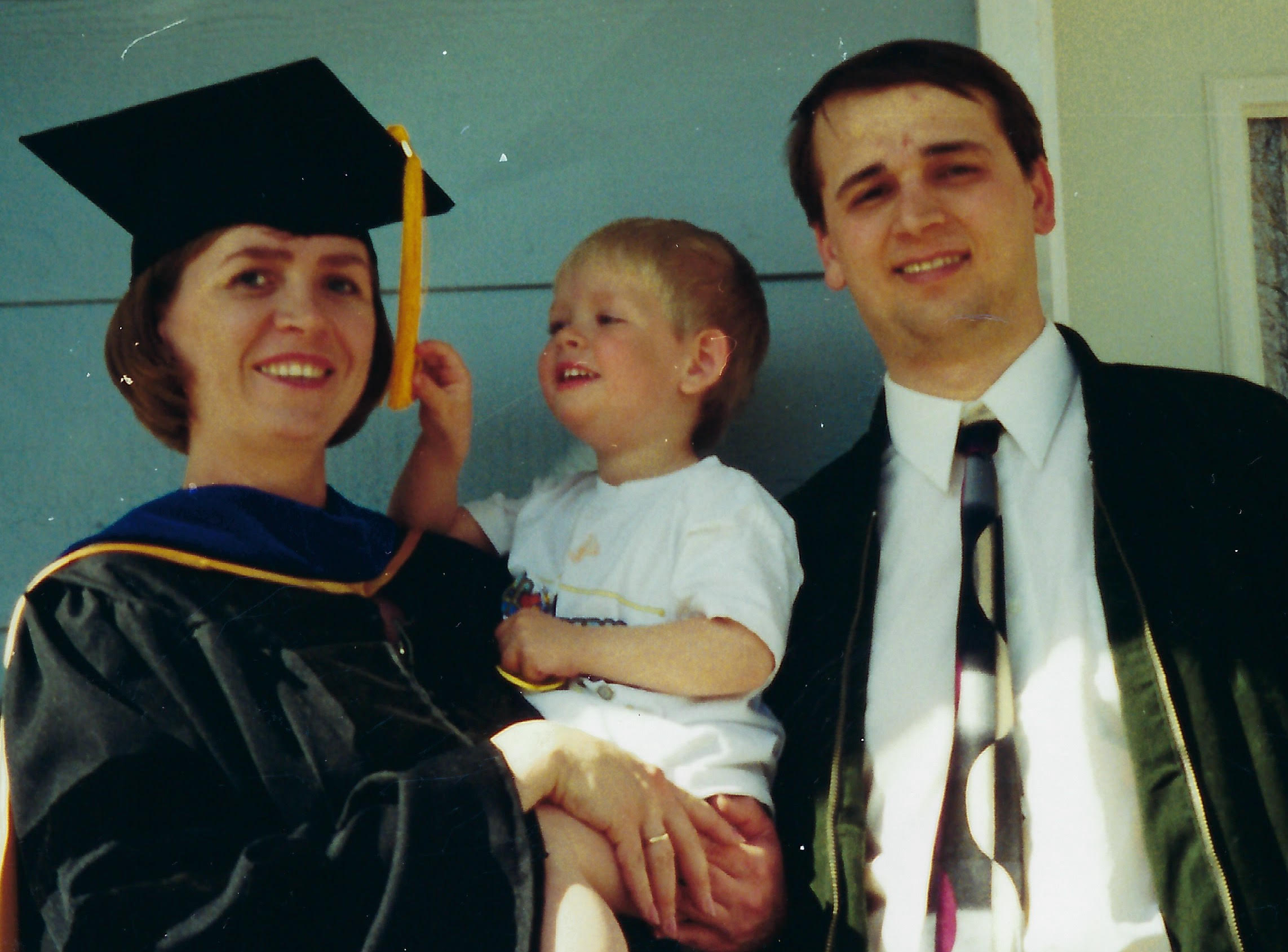 A woman in graduation regalia holds a young boy while standing next to a man in a tie.