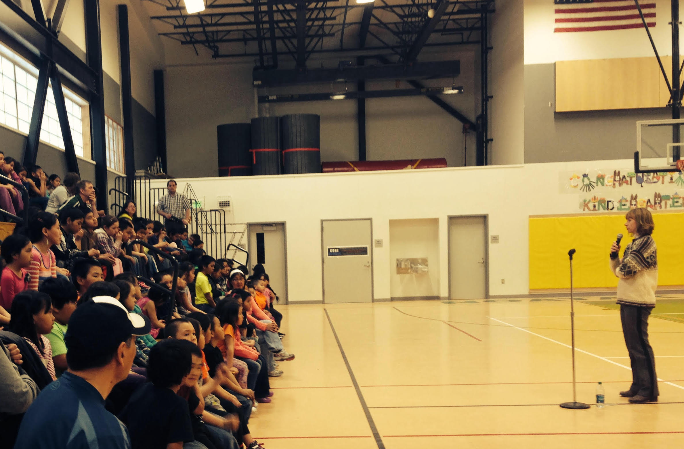 A woman speaks to young people on bleachers in a gymnasium.