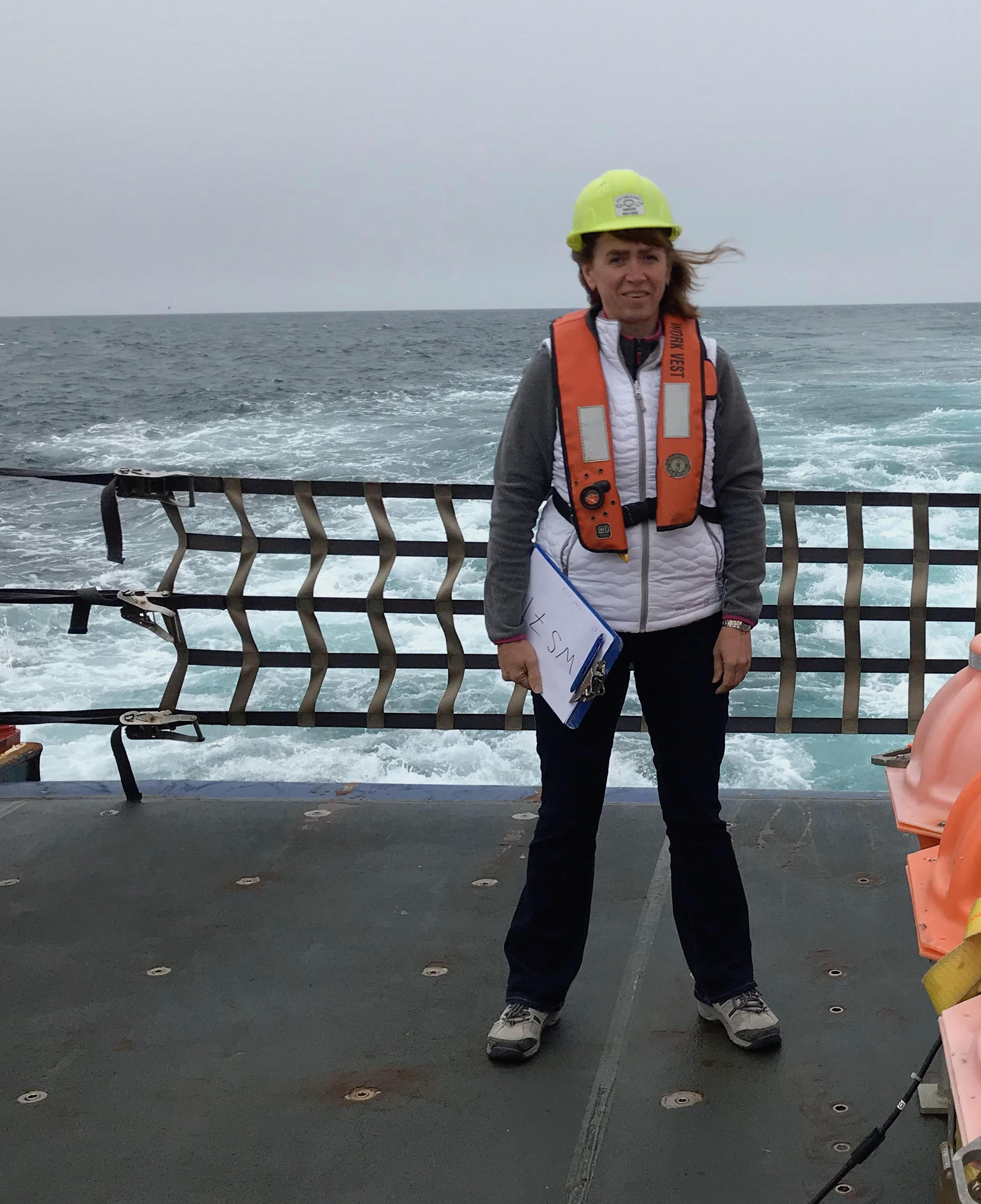 A woman stands on a ship deck while wearing a yellow hardhat and holding a clipboard.