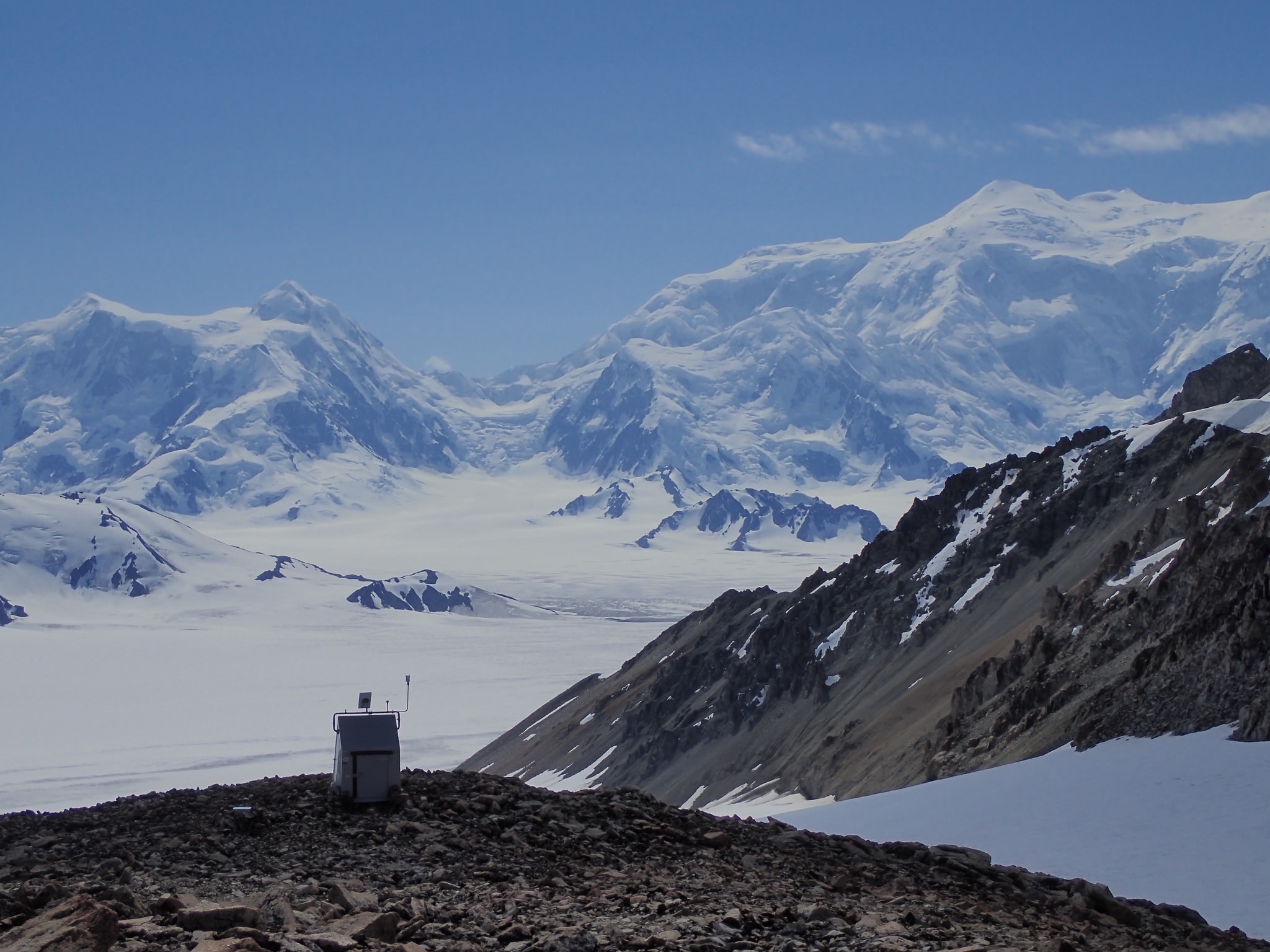 A seismic station sits on a rocky ridge with large ice- and snow-covered mountains in the distance on a sunny day.