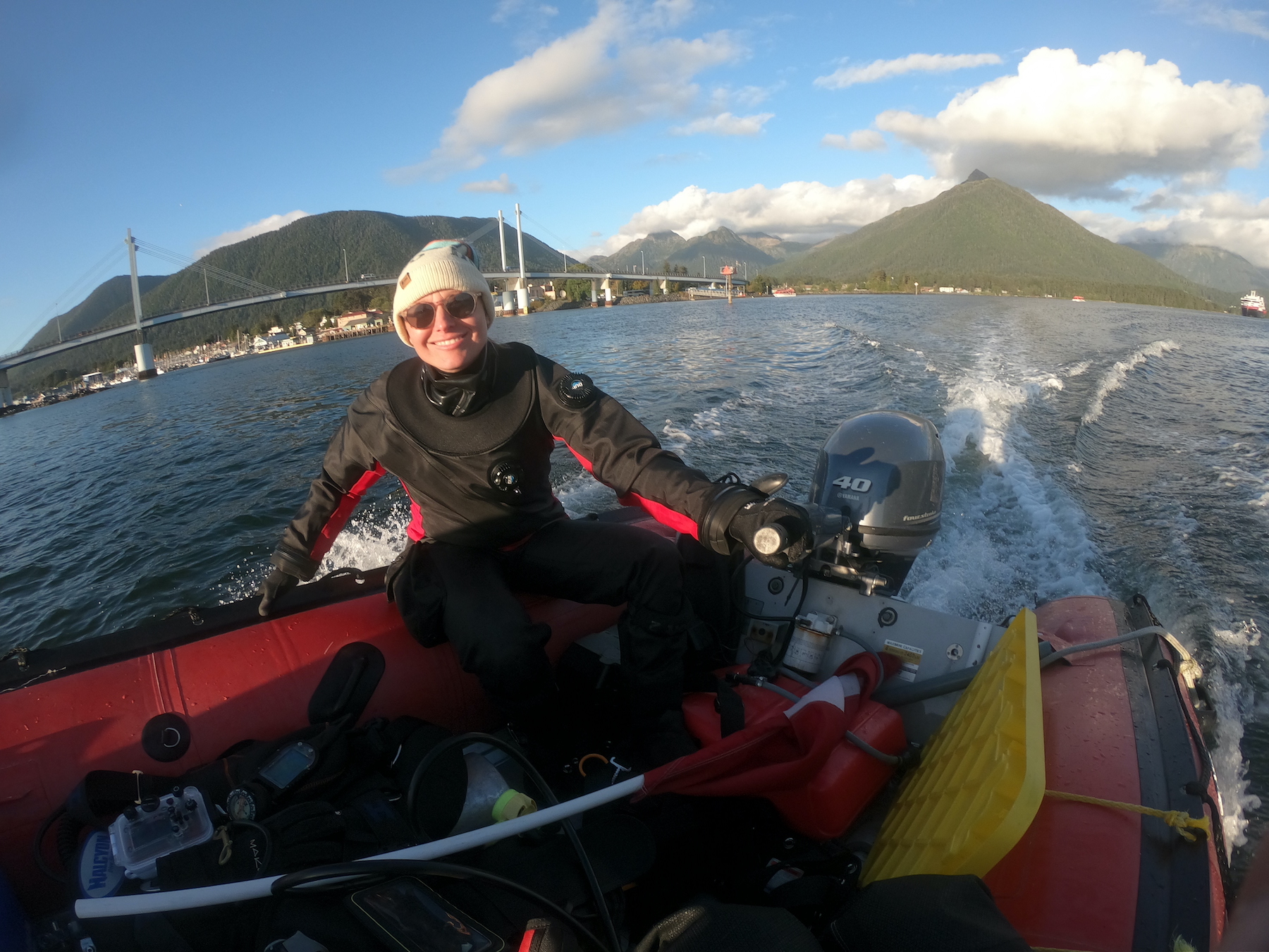 A woman in diving gear and a white knit cap sits on one tube of a red inflatable boat while driving the craft with a 40-horsepower outboard motor. A town, a marina, a suspension bridge, a ship and cloud-topped mountains are in the background on a mostly sunny day.