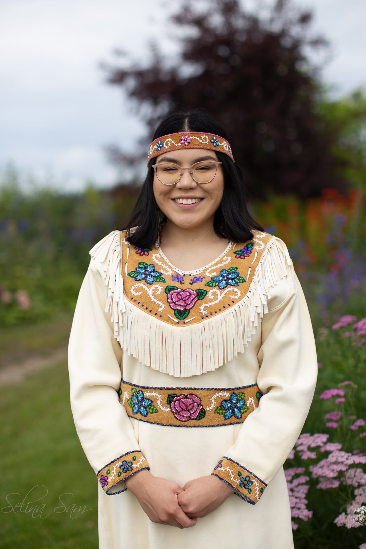 Laura Ekada stands outdoors among wildflowers in bloom in her traditional Athabascan regalia.