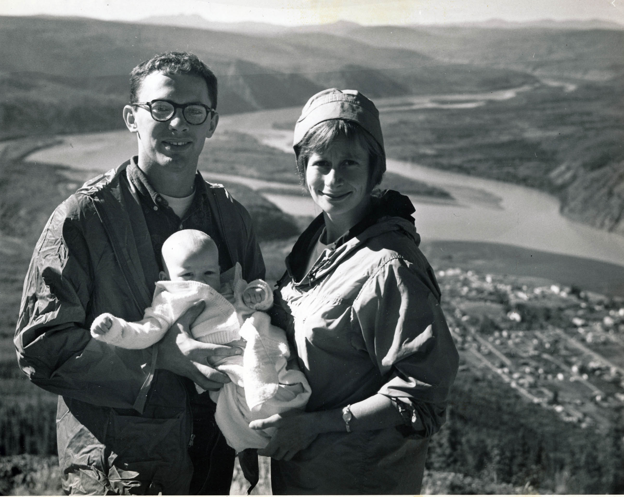 A black and white photo of a man and woman hold a baby while standing atop a hill with a view of Dawson City, in Canada's Yukon, behind them.
