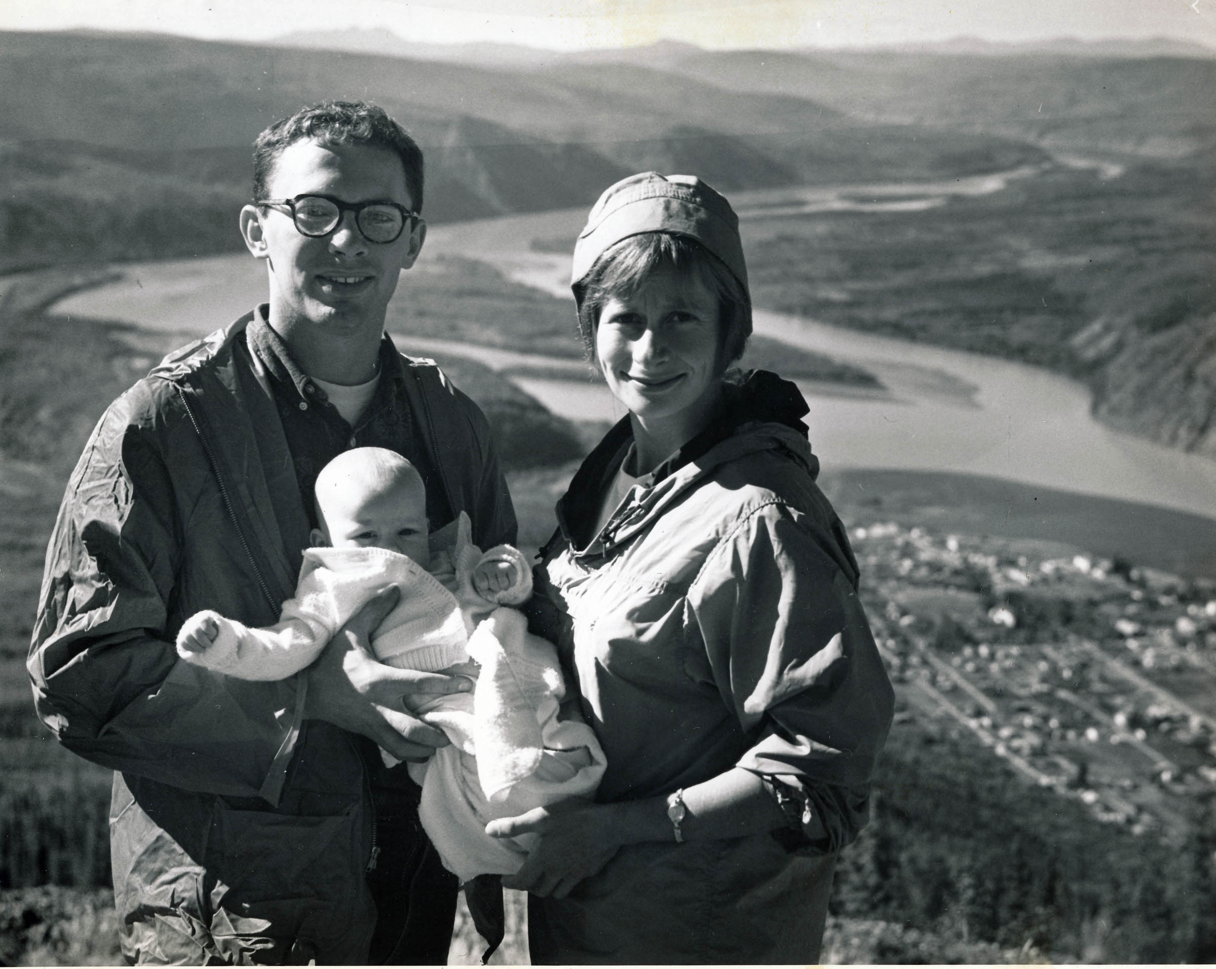 A man and a woman stand on a promontory holding a baby. Behind them, far below, is a town and river winding between high hills. The image is black and white.