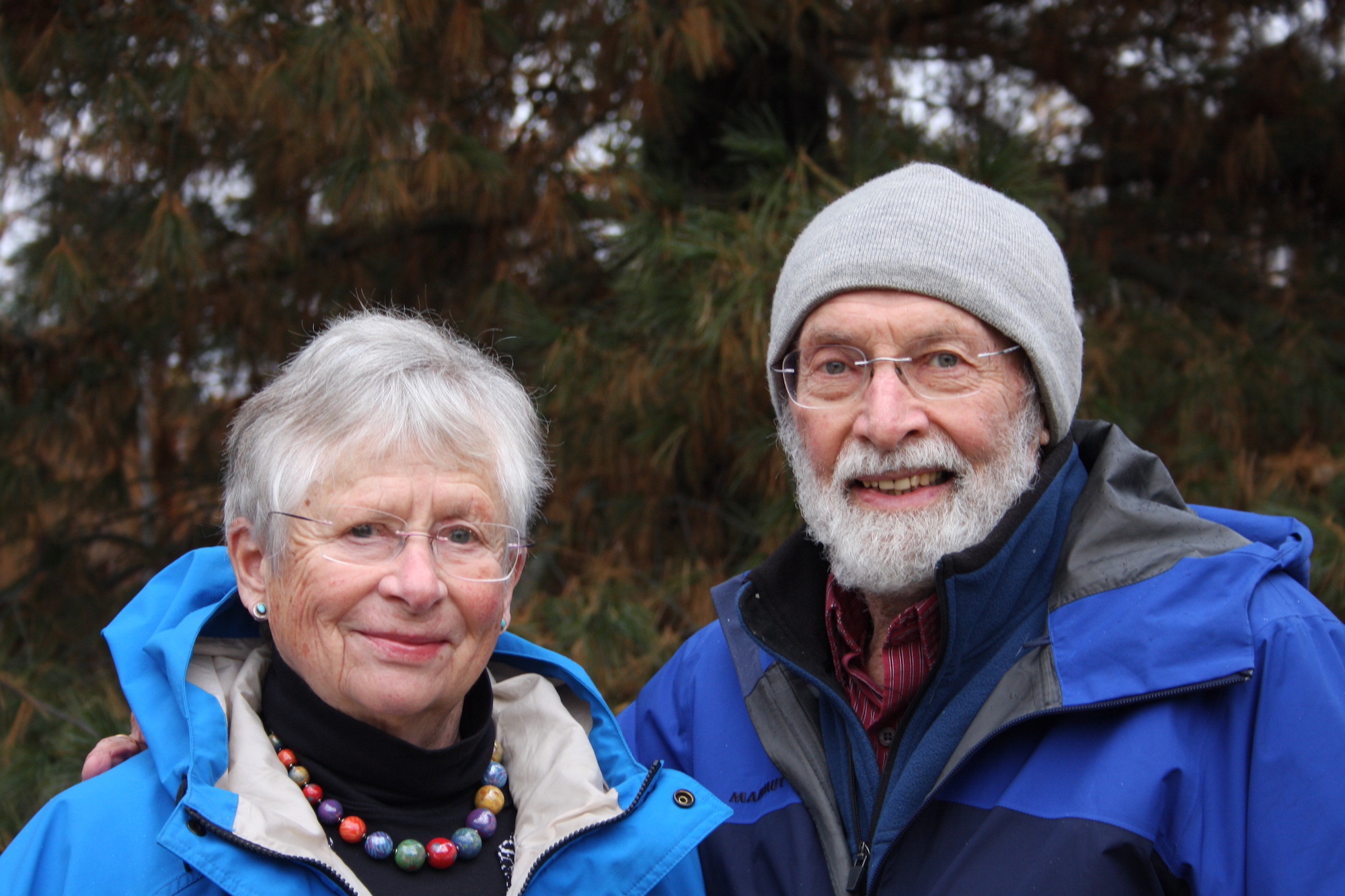 A woman and a man, both with gray hair and glasses and wearing blue raincoats, smile at the photographer. The man has a beard and is wearing a knit cap.