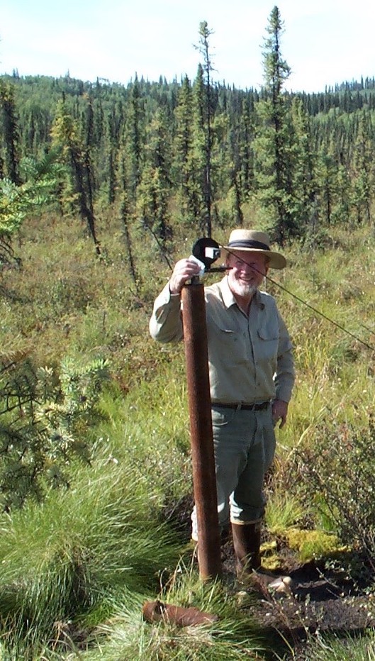 On a sunny summer day, a man in a brimmed hat, khaki clothing and rubber boots stands next to a rusty iron pipe topped with a scientific instrument  and surrounded by a forest of Alaska's small black spruce trees.