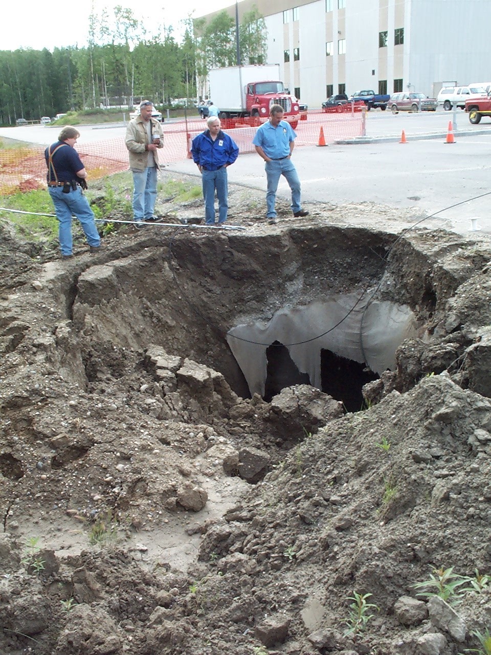 Several men stand around a large sinkhole, with a parking lot and a university building in the background.