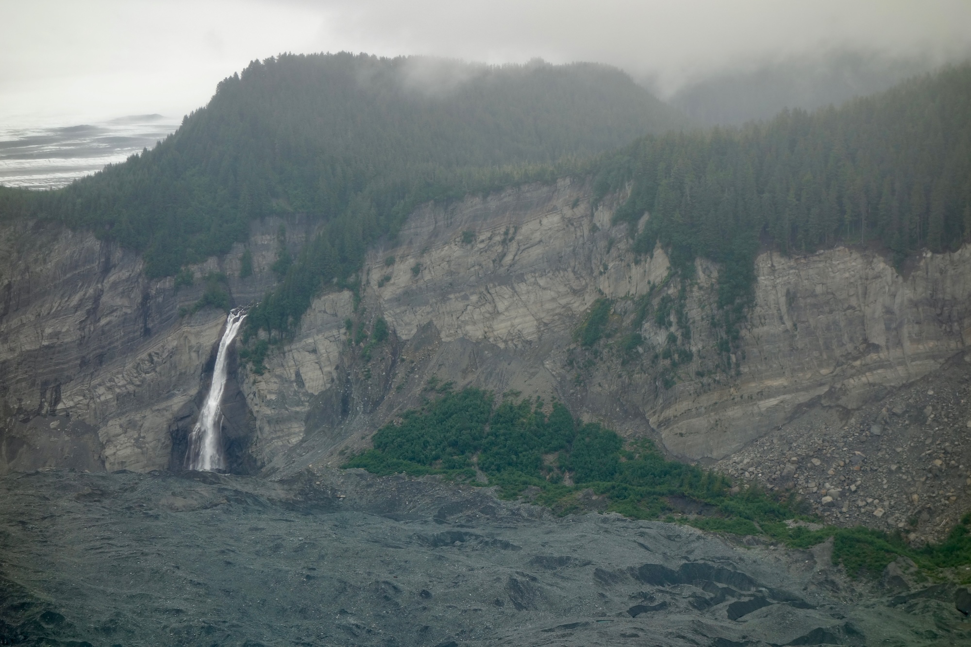 A waterfall falls from the top of a cliff that is topped with evergreens.