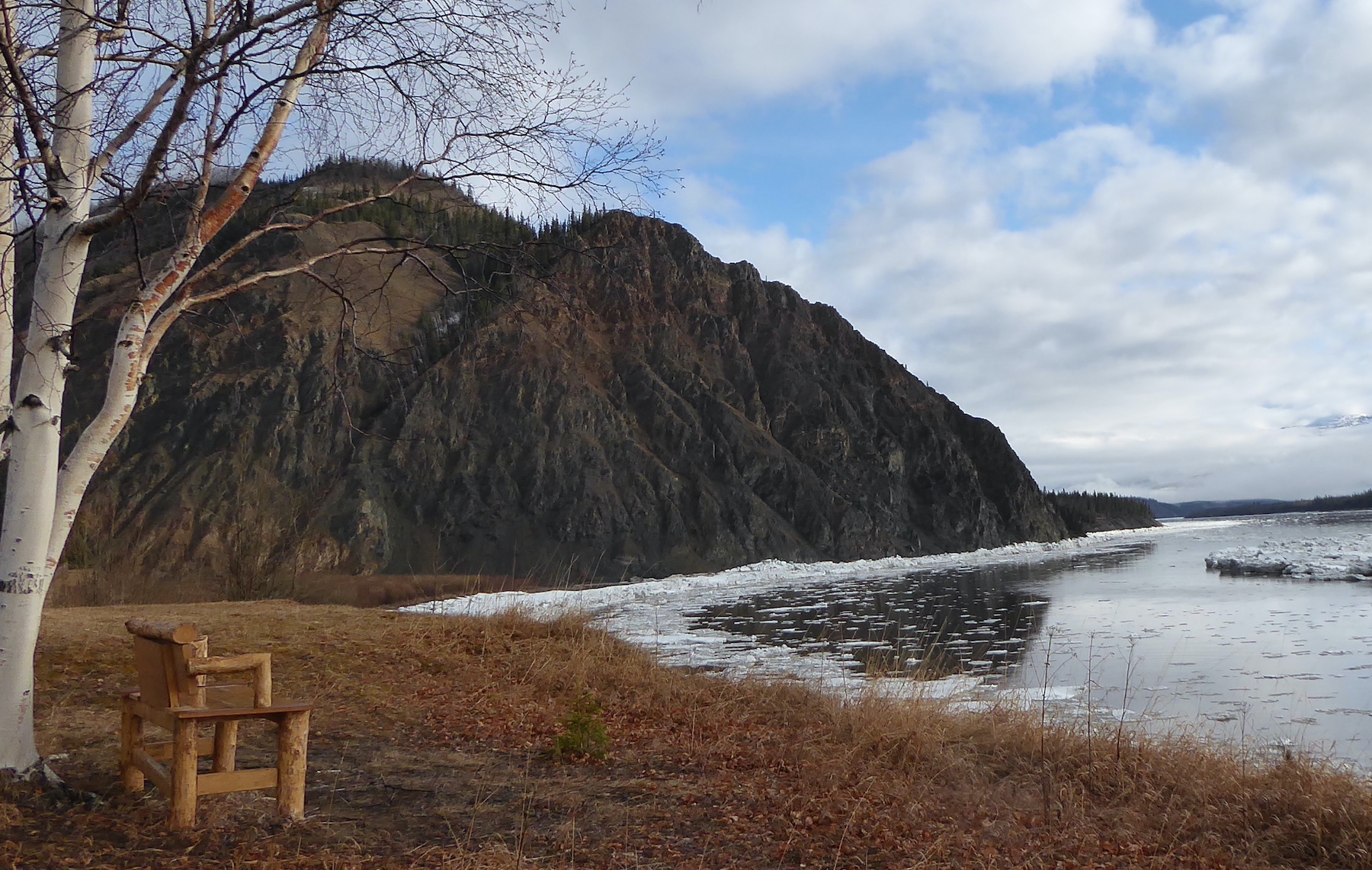 Crushed river ice lies on the bank below a large cliff. In the foreground, a bench sits under a birch tree overlooking the river.
