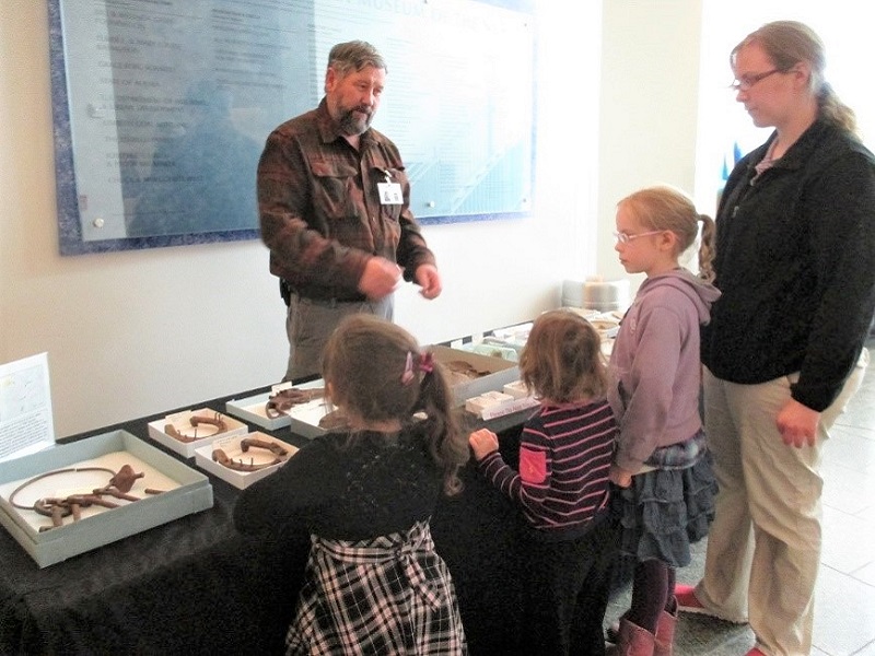A bearded man in a flannel shirt stands behind a table loaded with archaeological artifacts. He is talking to three children and an adult woman.