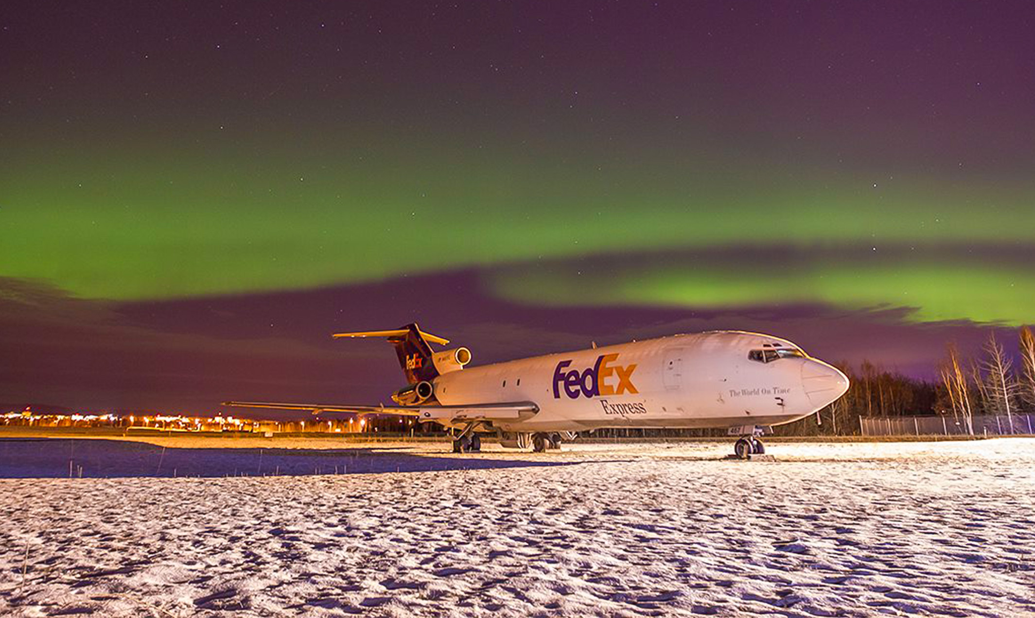 A Boeing 727 jet with a FedEx logo sits on a snowy runway under the northern lights.
