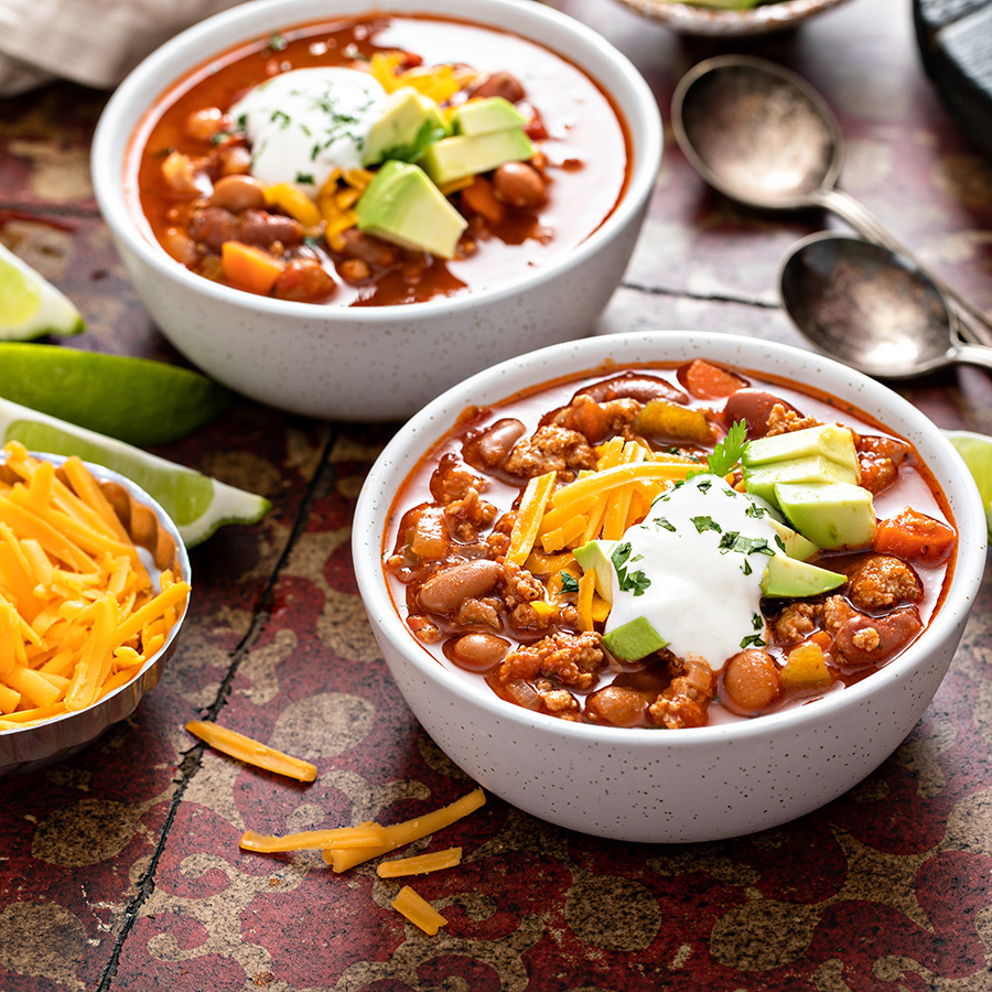 Two bowls of chili and two spoons on a table with cheese and limes nearby.
