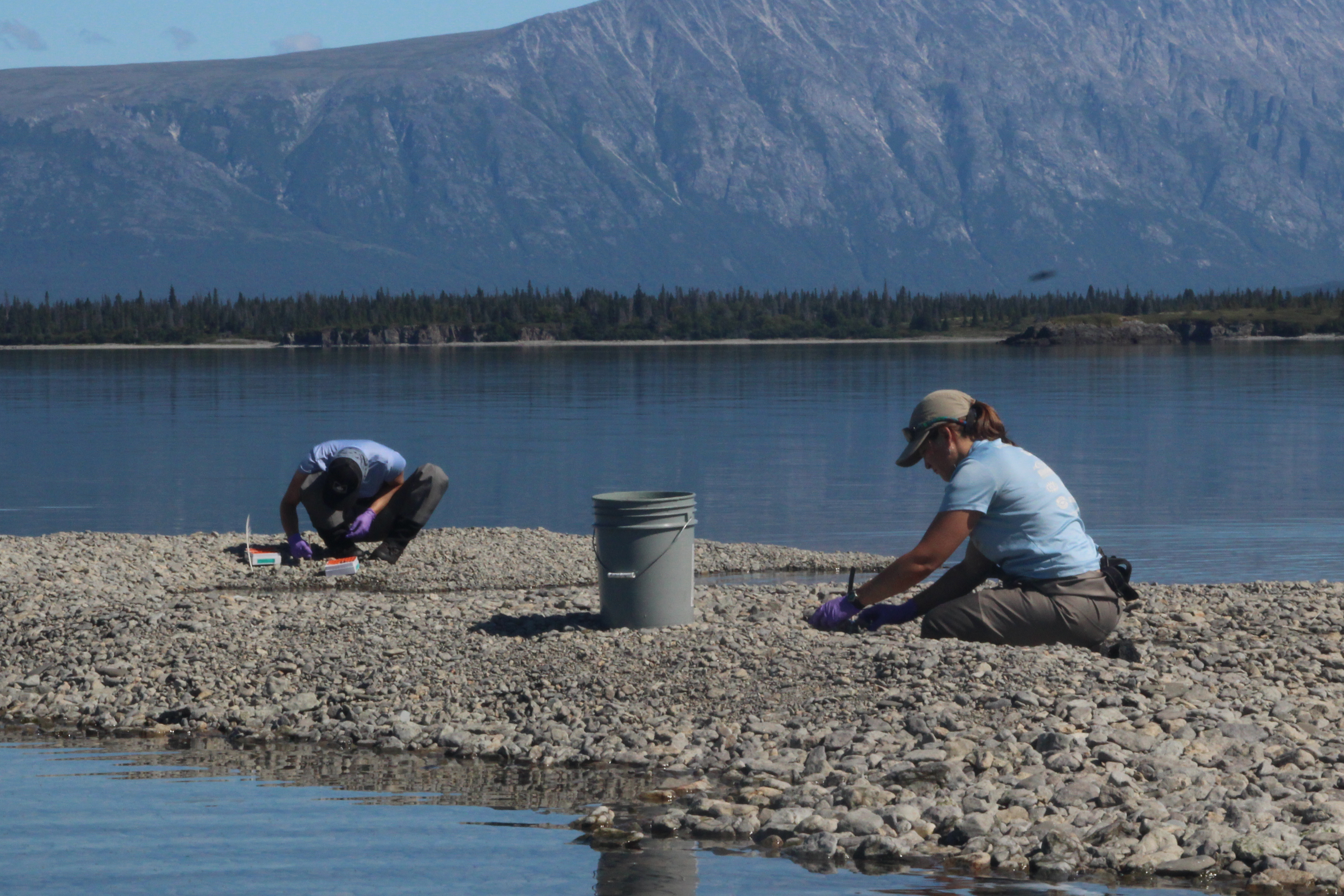 two people crouched on a rocky shore with a bucket and mountains in the background