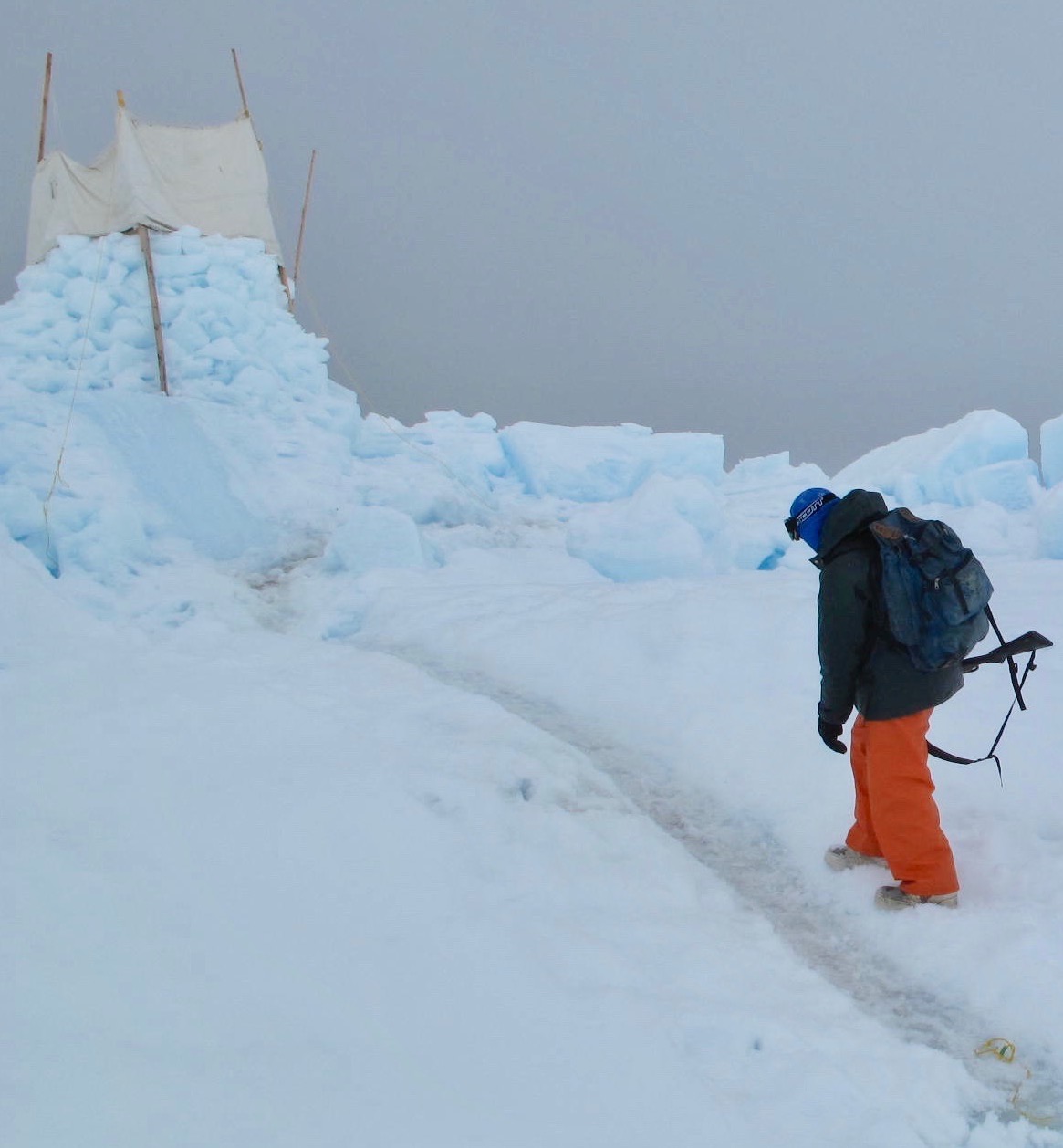 A man in winter gear carrying a rifle climbs a ridge of ice with a snow-block structure at the top. A canvas sheet rises above the snow blocks.