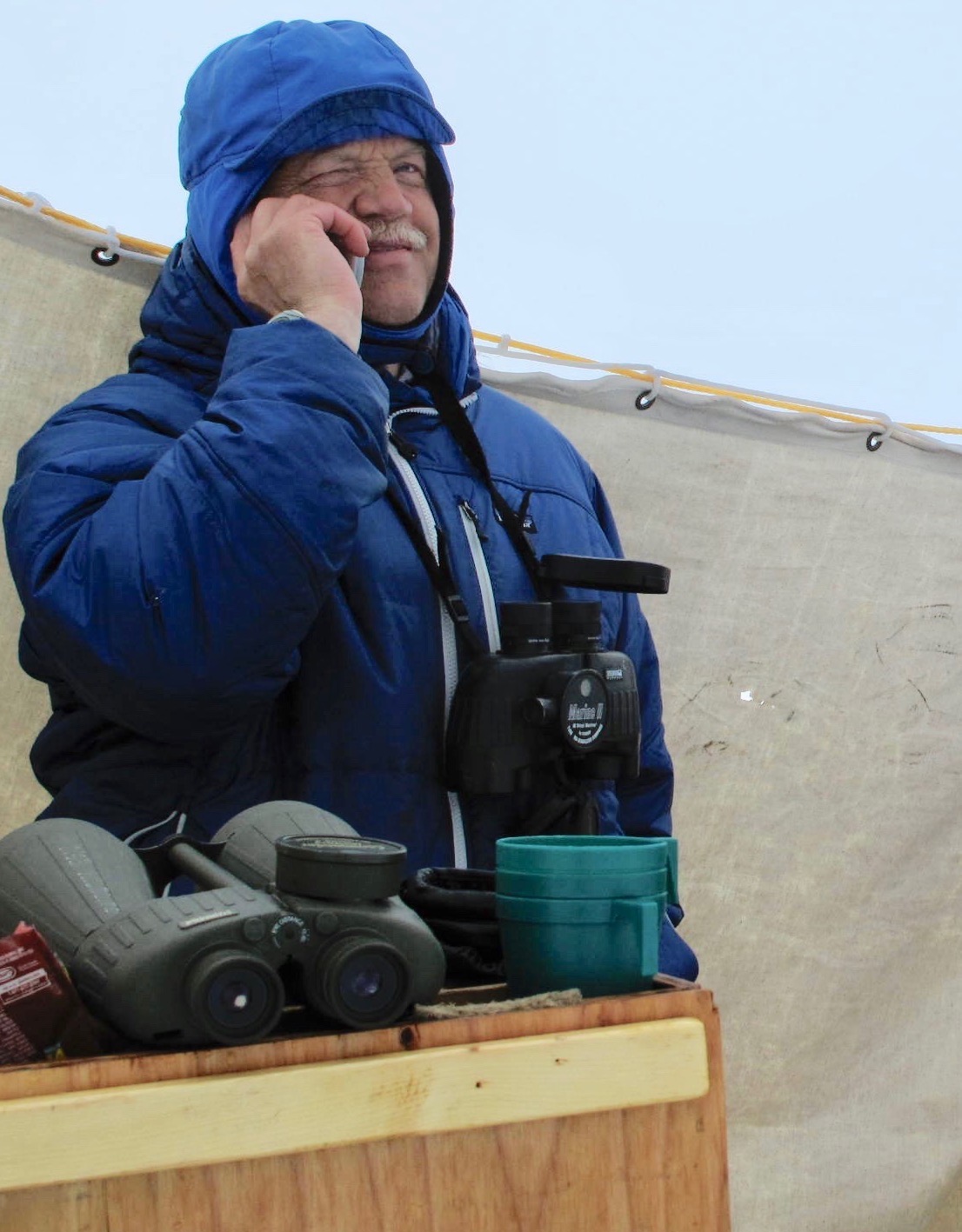 A man in a blue winter hat and coat speaks on a cell phone. A set of binoculars hangs around his neck and another sits on a plywood box in the foreground, along with a stack of green plastic cups. A white canvas sheet strung along a rope forms a backdrop. 