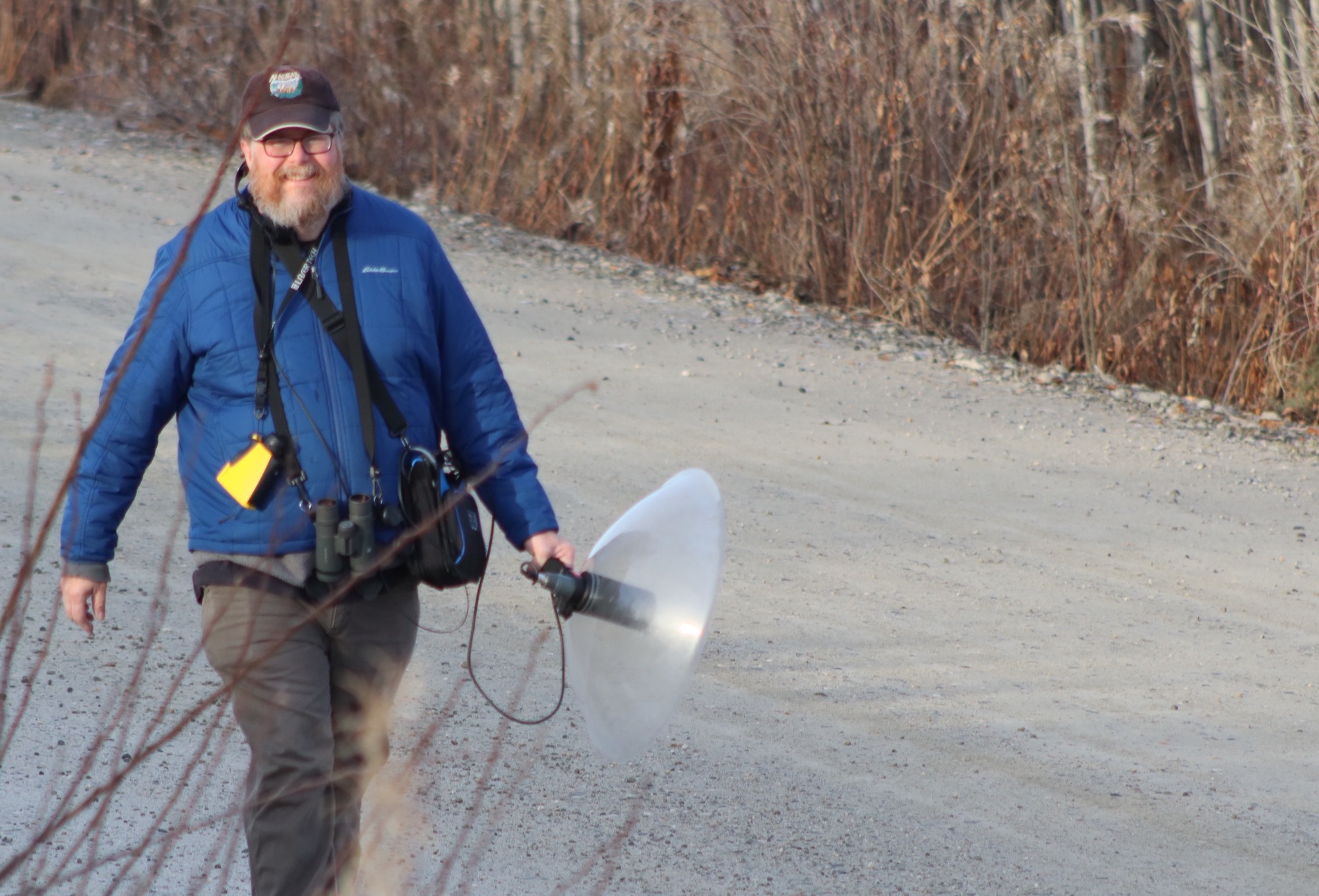 A man in a blue insulated jacket and ball cap walks down a gravel road lined with fall-season brush while carrying a microphone with a dish receiver.