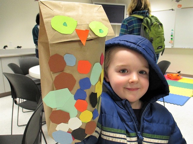 A young child in a blue jacket with hood smiles and holds up an owl crafted from a paper bag and colored construction paper.