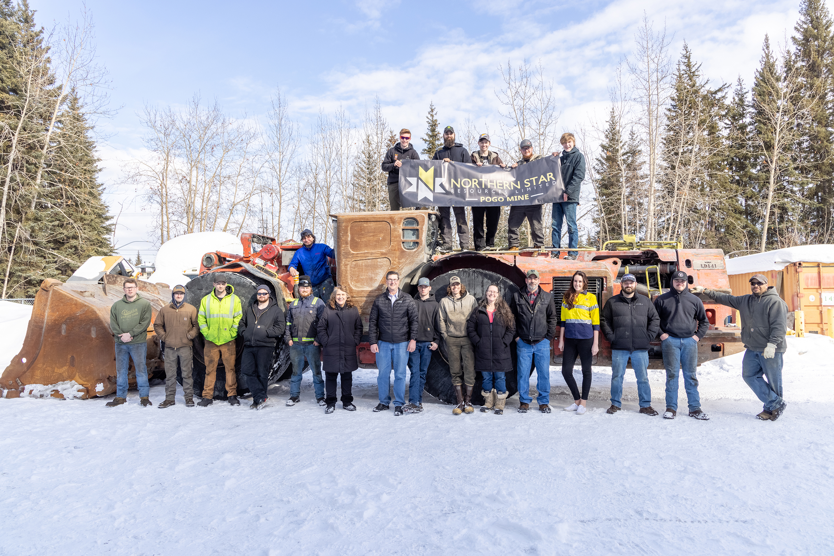 A group of more than a dozen people pose in front of a piece of heavy equipment.