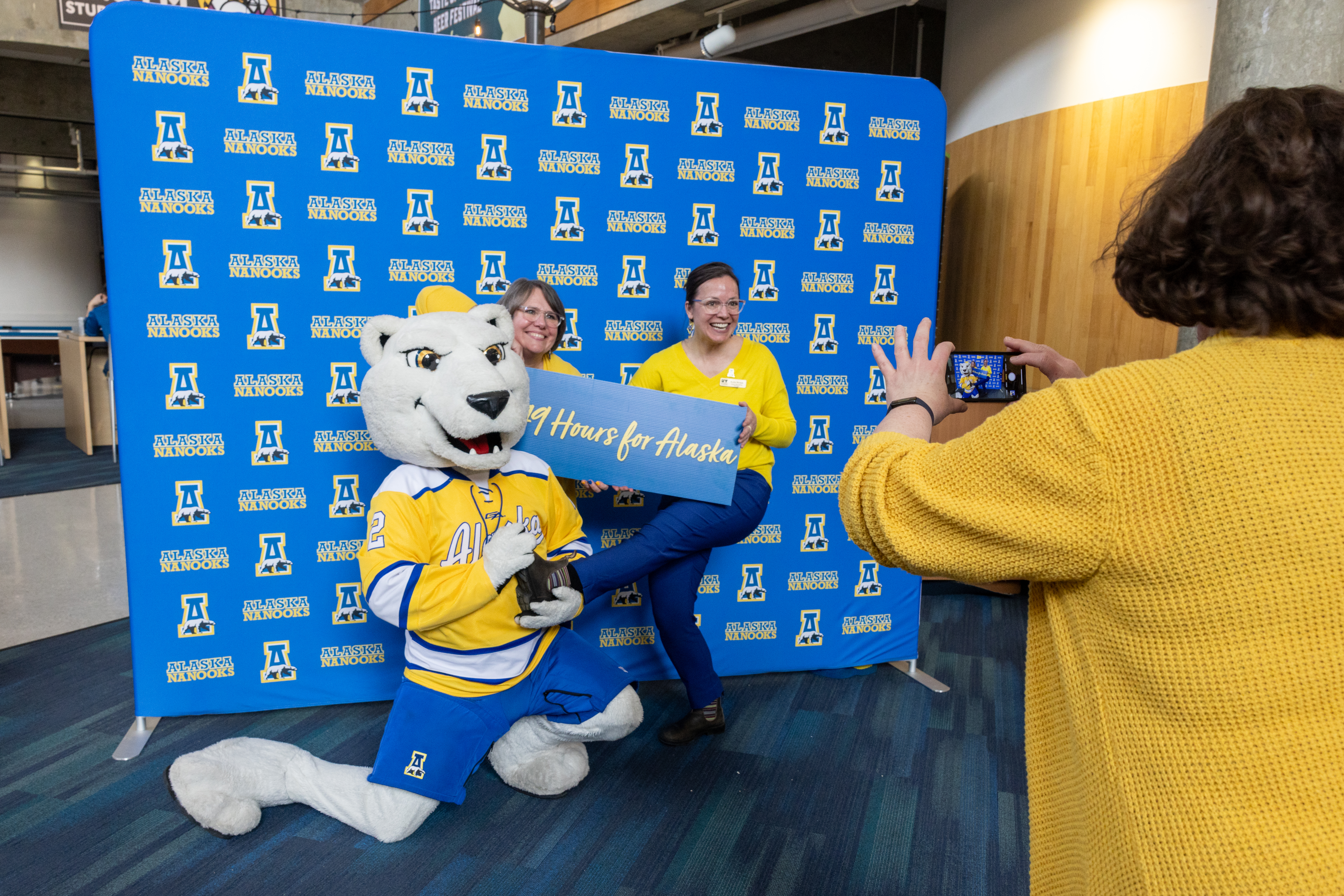 a polar bear mascot and two women hold a sign saying "#49HoursForAlaska" in front of a blue backdrop while someone takes their picture.