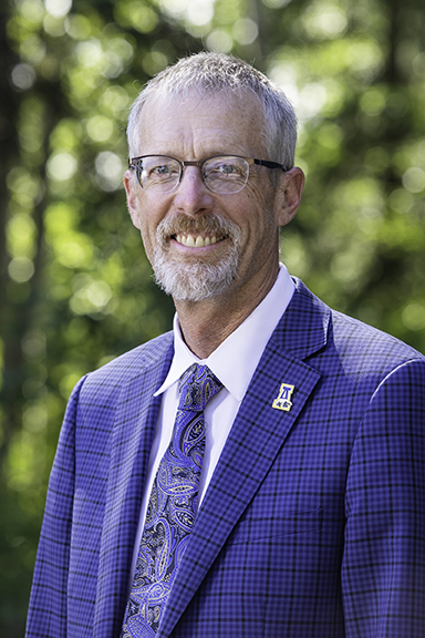 Head and shoulders photo of UAF Chancellor Dan White outdoors wearing a blue and black plaid blazer with a UAF athletics pin over a white collared shirt and blue paisley necktie with yellow accents.