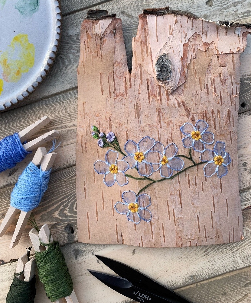 Photo of forget-me-not flowers hand embroidered onto birch bark with colored thread wrapped around clothespins and a pair of scissors next to the art piece.