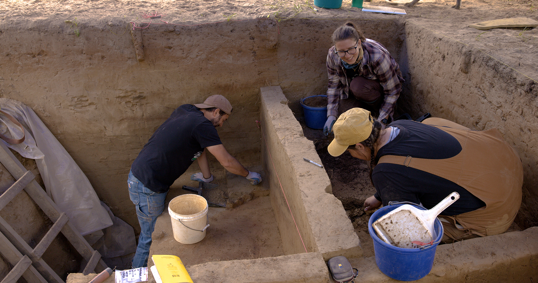 three people work in a square hole in the ground with various buckets, tools and ladders